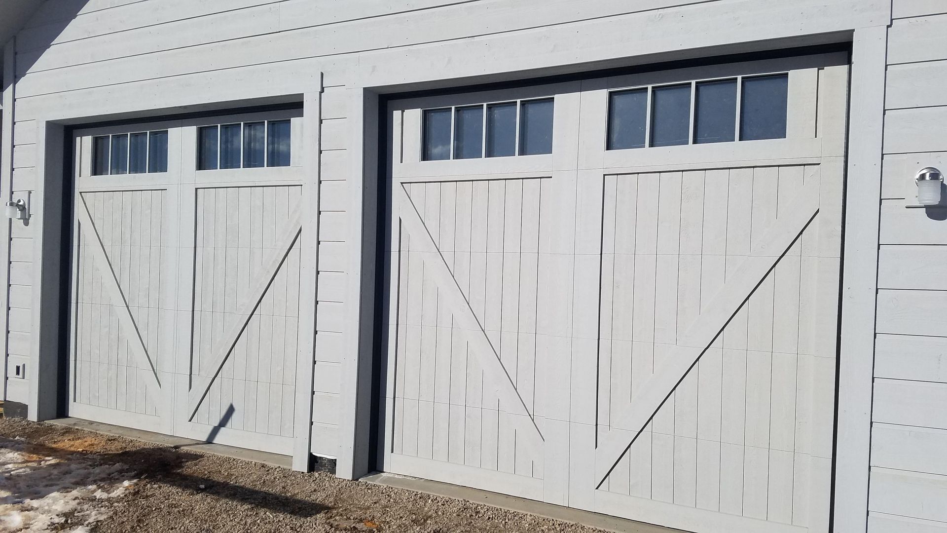 Two white garage doors with windows and diagonal trim on a light gray house.