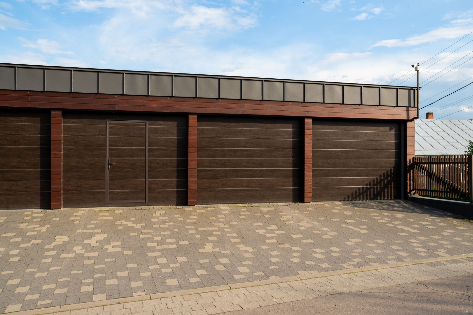 Brown garage doors with a brown roof and brick driveway against a blue sky.