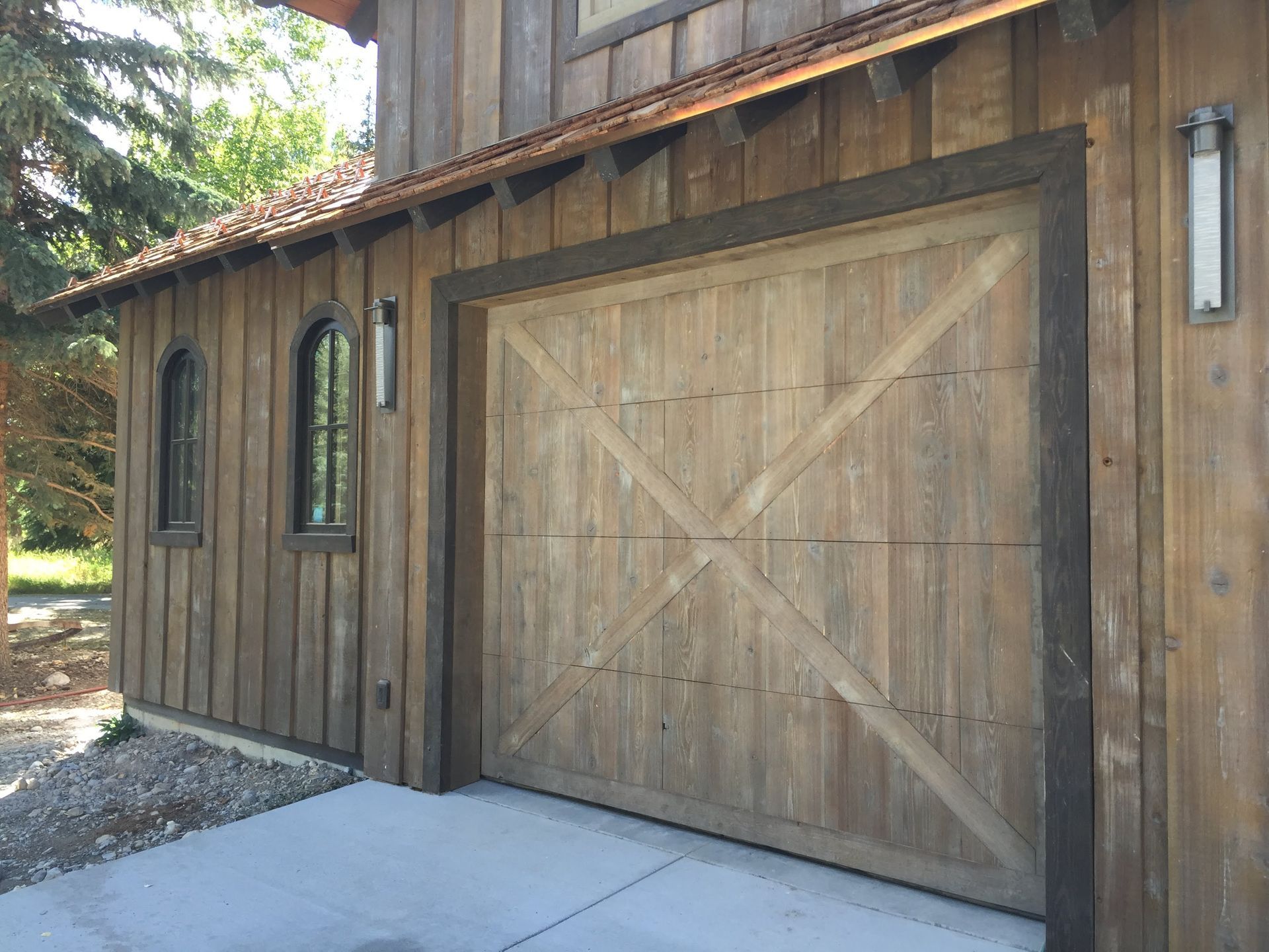 Wooden garage door with X-patterned trim on rustic wood-clad building. Two sconces and arched window on left.