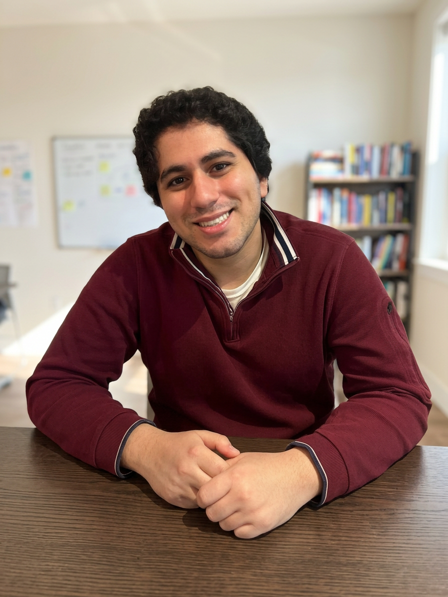 Omar Shohoud is smiling in a maroon quarter-zip sweater, and sits at a wooden table in a classroom setting with a bookshelf behind.