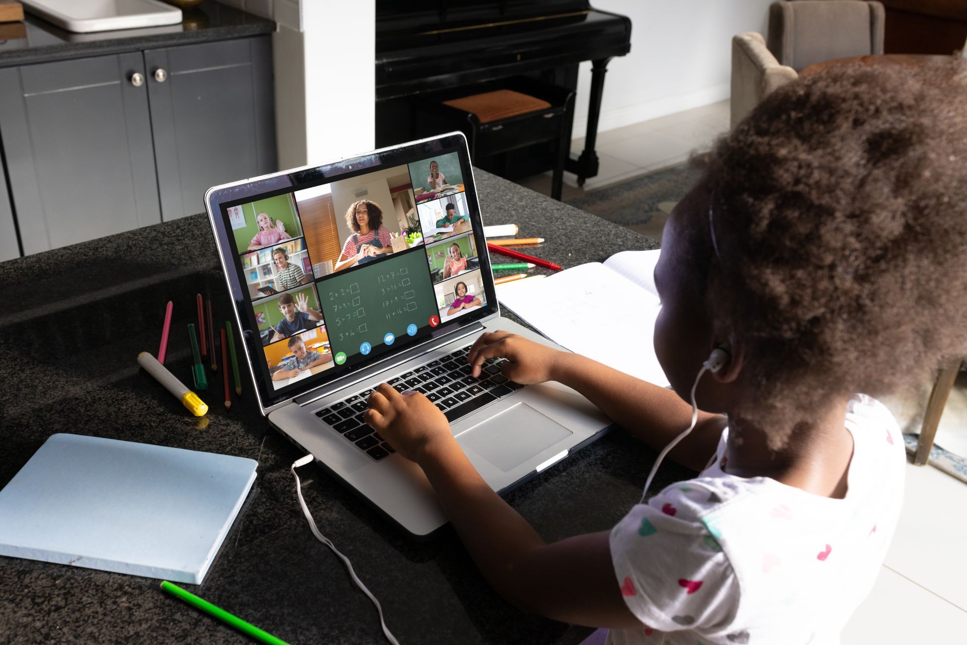 A little girl sits in front of a laptop, attending a virtual maths class with eight other students and the teacher.