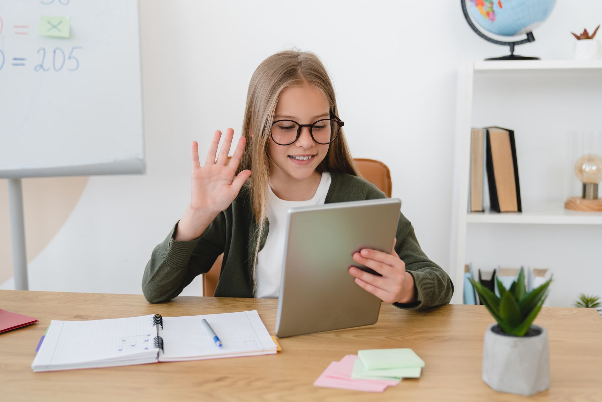 A student at a desk waves at a tablet screen while taking a virtual class with an open notebook and a small plant nearby.