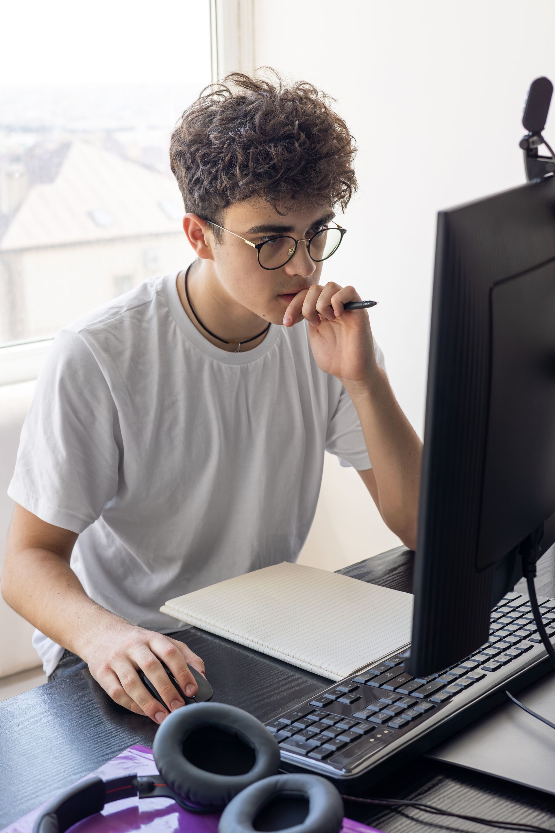 A person with curly hair wearing glasses works at a desk, looking at a computer screen while holding a pen.