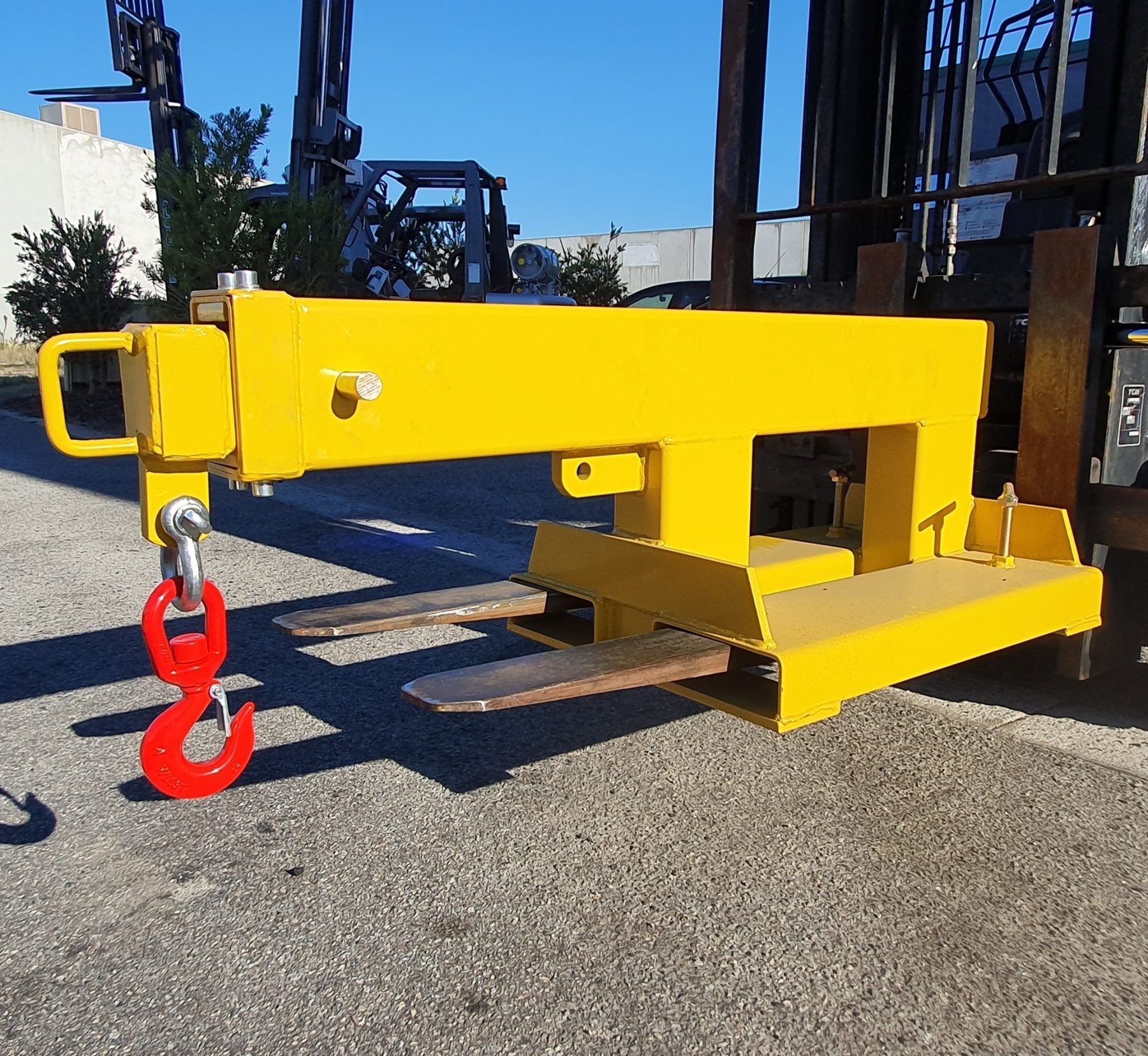 Yellow forklift attachment with a red lifting hook and two forks; outdoors.