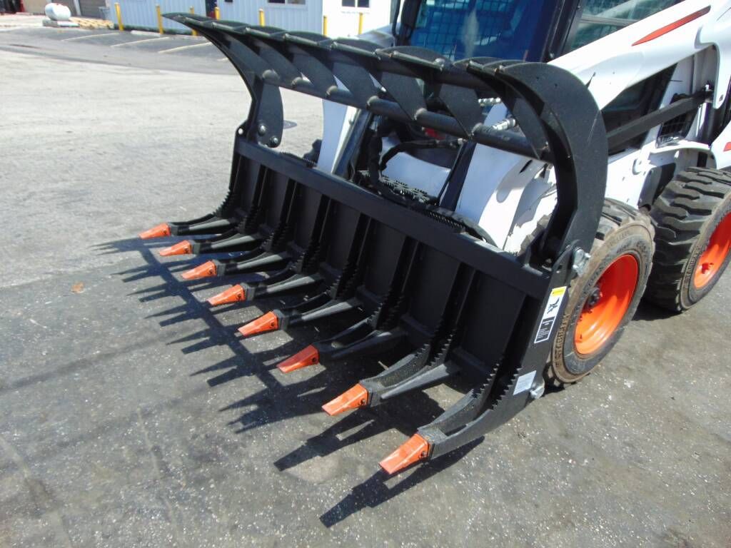 Bobcat skid-steer with grapple attachment, black tines, orange tips, on a paved surface.