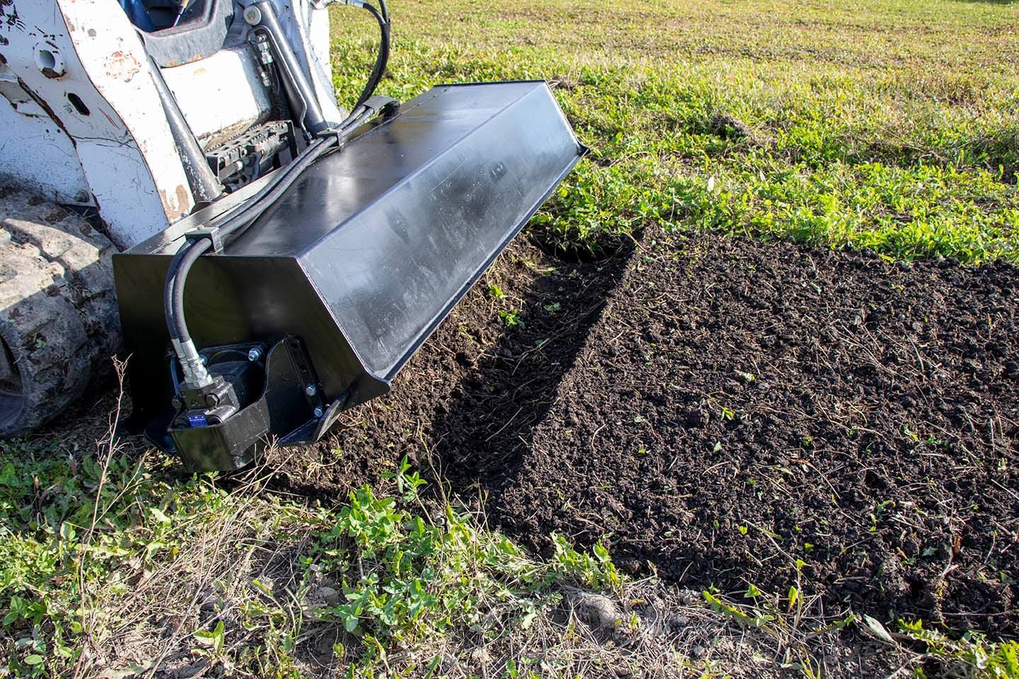 Skid steer with black attachment tilling a dark, freshly-turned patch of soil in a grassy field.