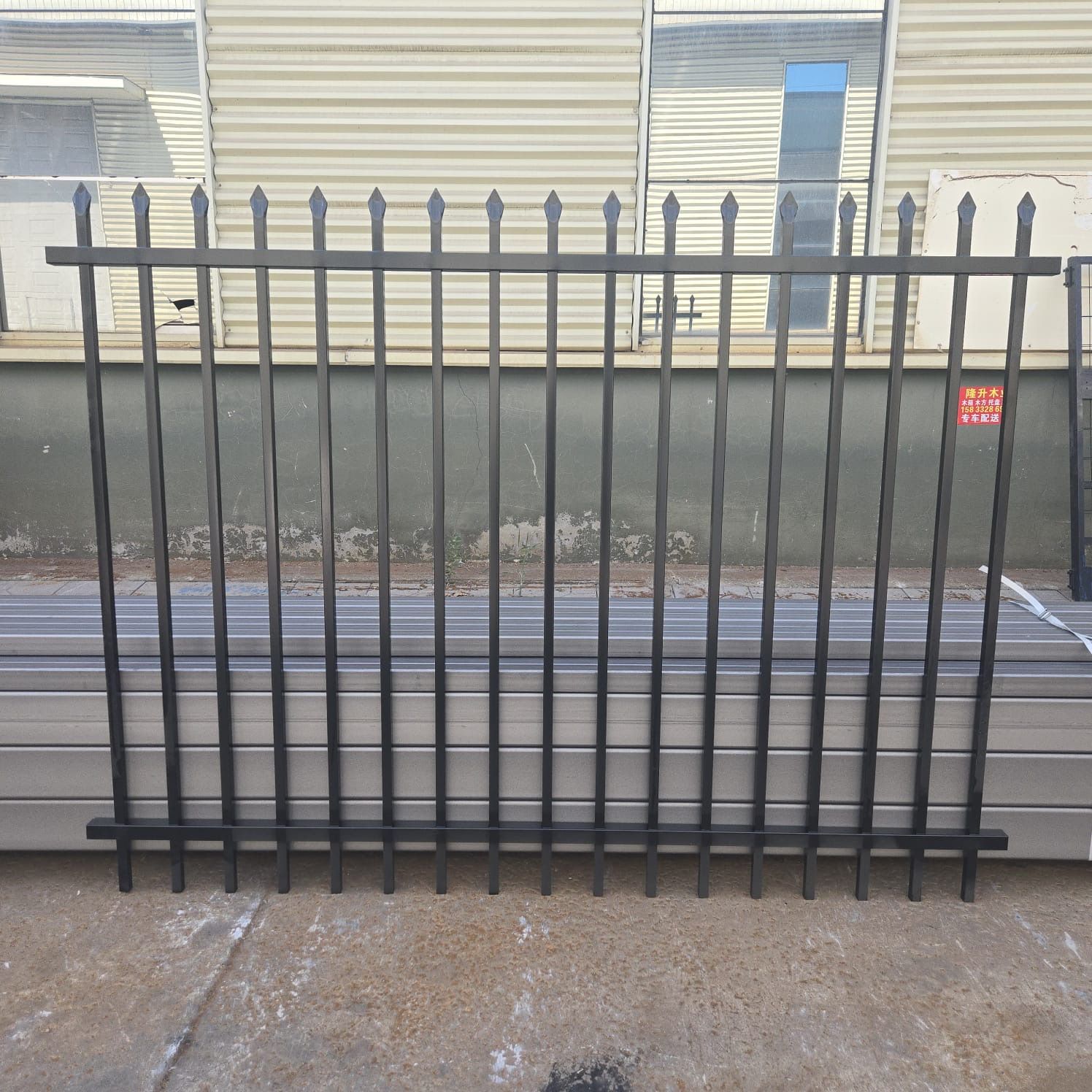 Black metal fence section with pointed top pickets, set against a corrugated metal building.