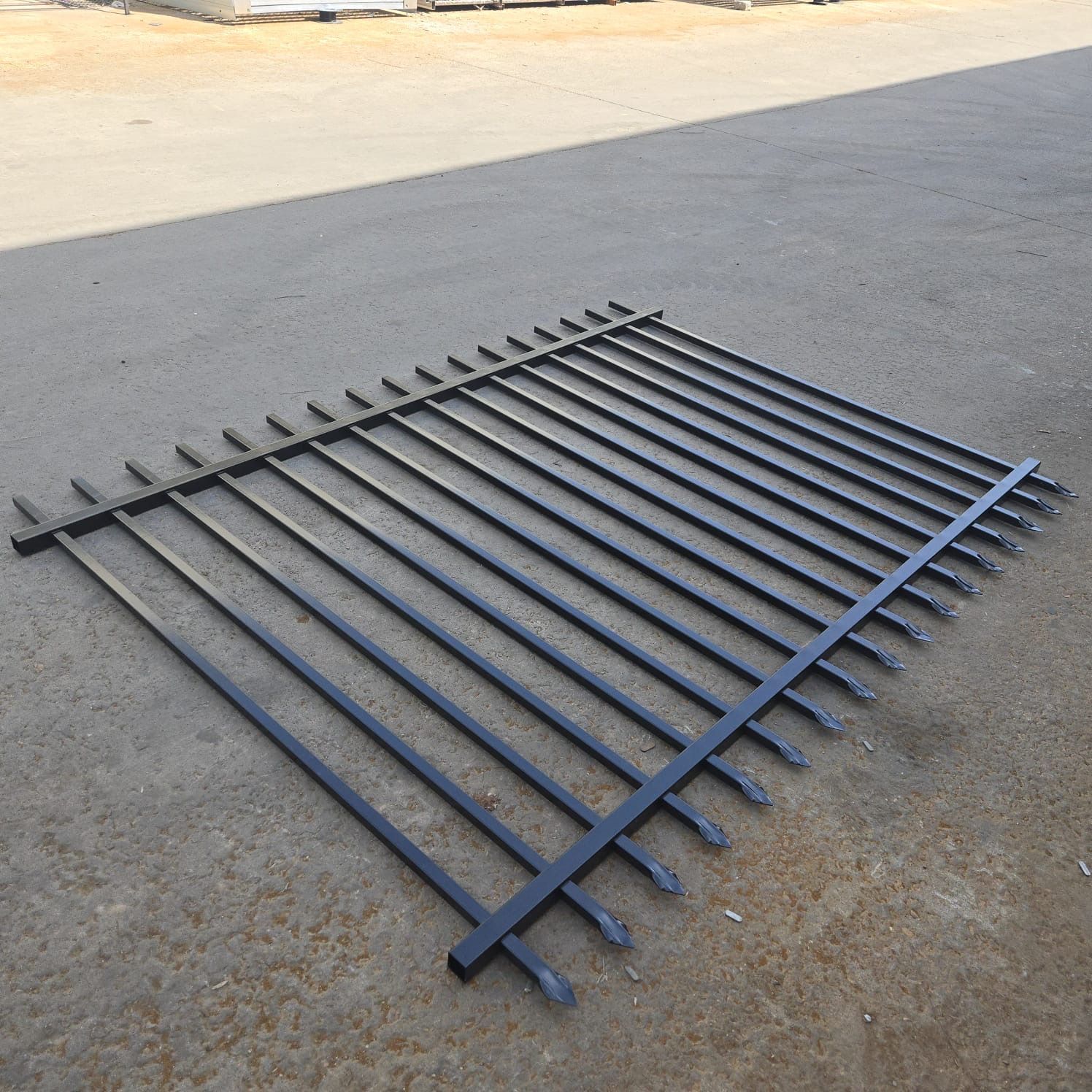 Black metal fence section with pointed top pickets, set against a corrugated metal building.
