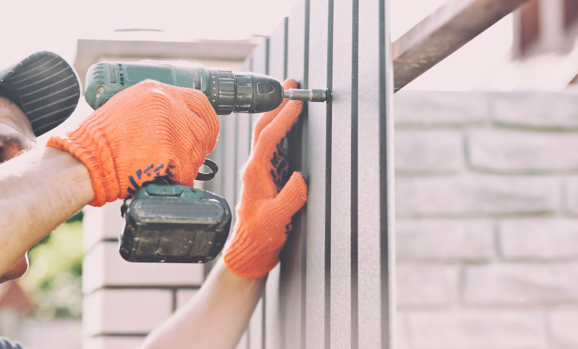 Person in orange work gloves using a drill to screw into a fence.