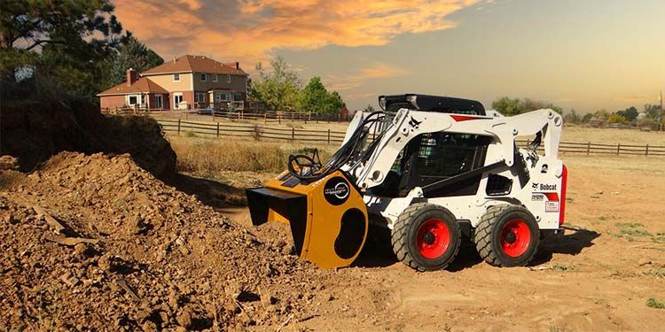 White Bobcat skid-steer loader with a soil compactor attachment on a construction site next to a home.