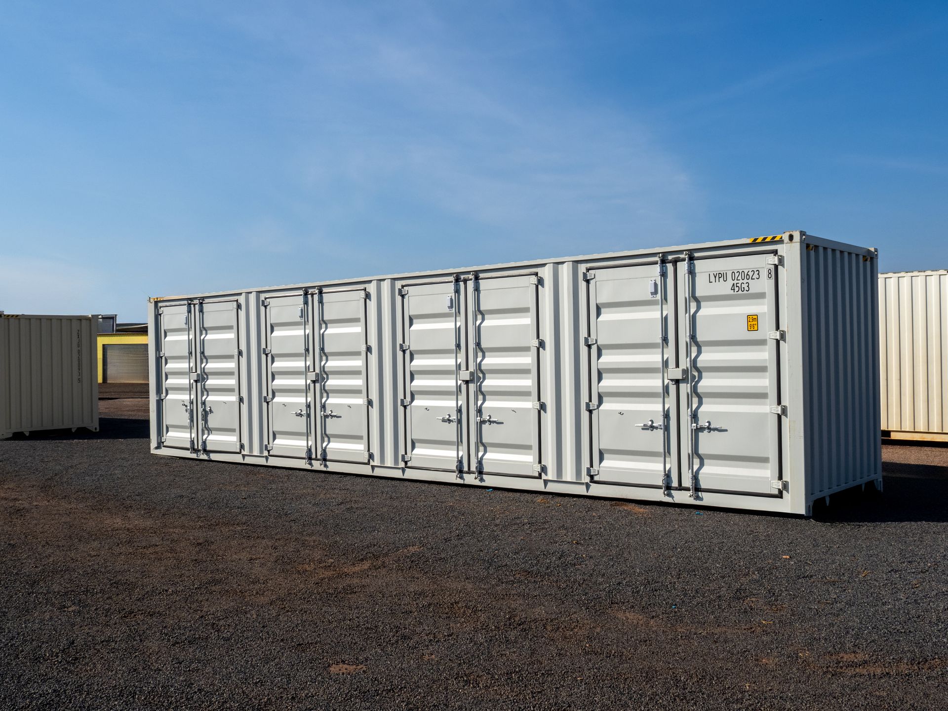 Five stacked, light-gray shipping containers on a gravel lot under a blue sky.