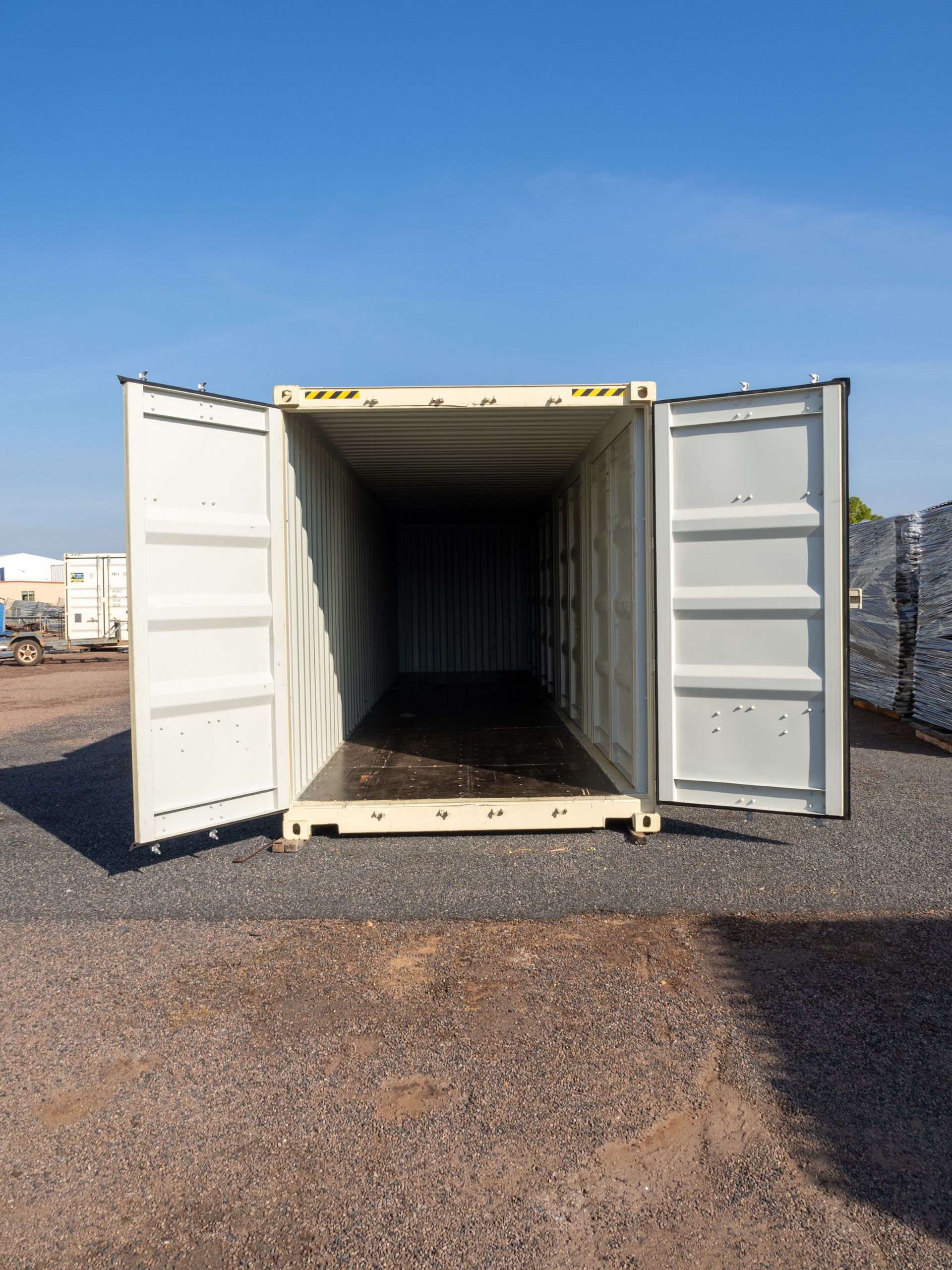 Empty shipping container with doors open on gravel under blue sky.
