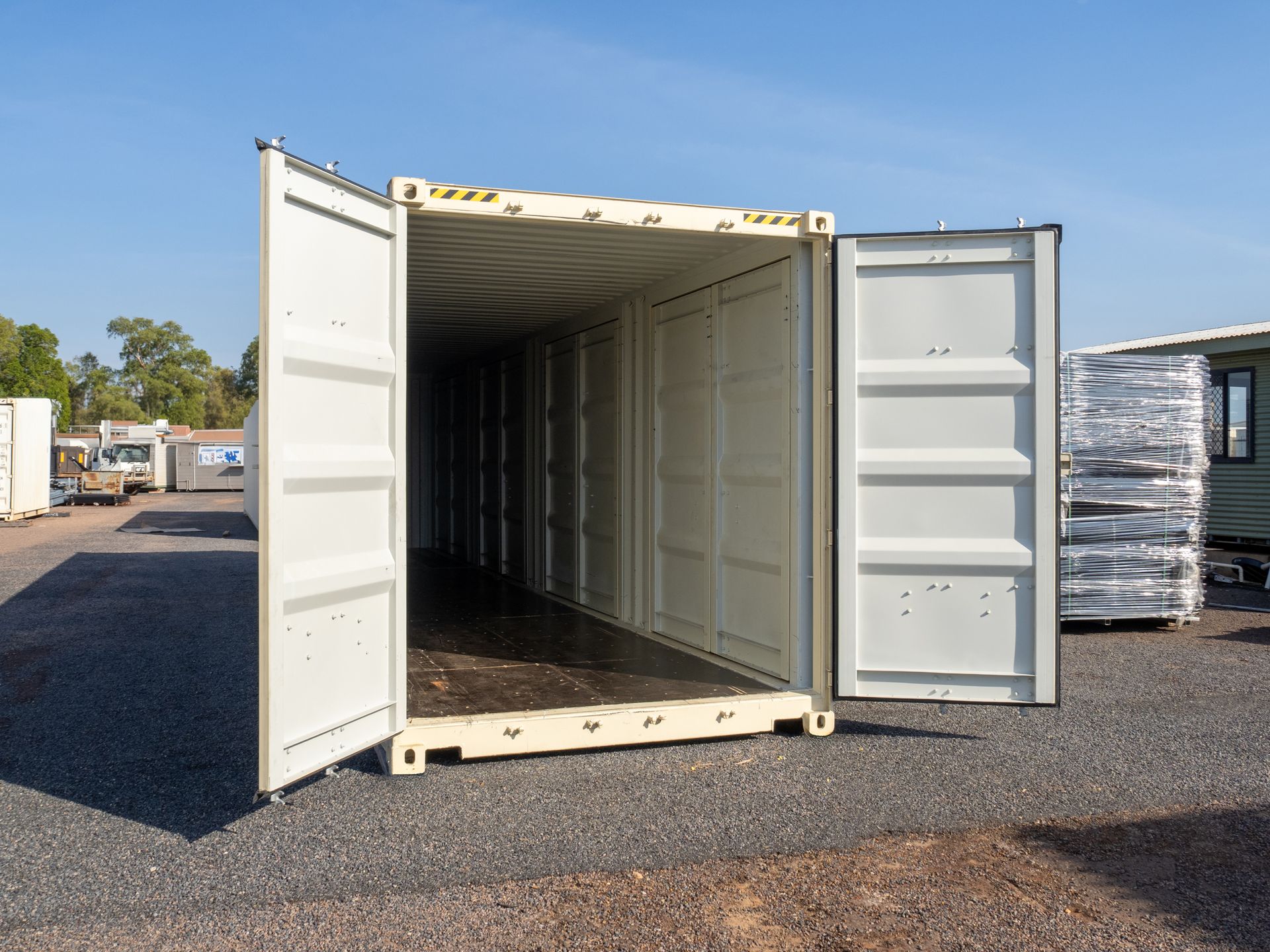 Empty beige shipping container with doors open on gravel surface.