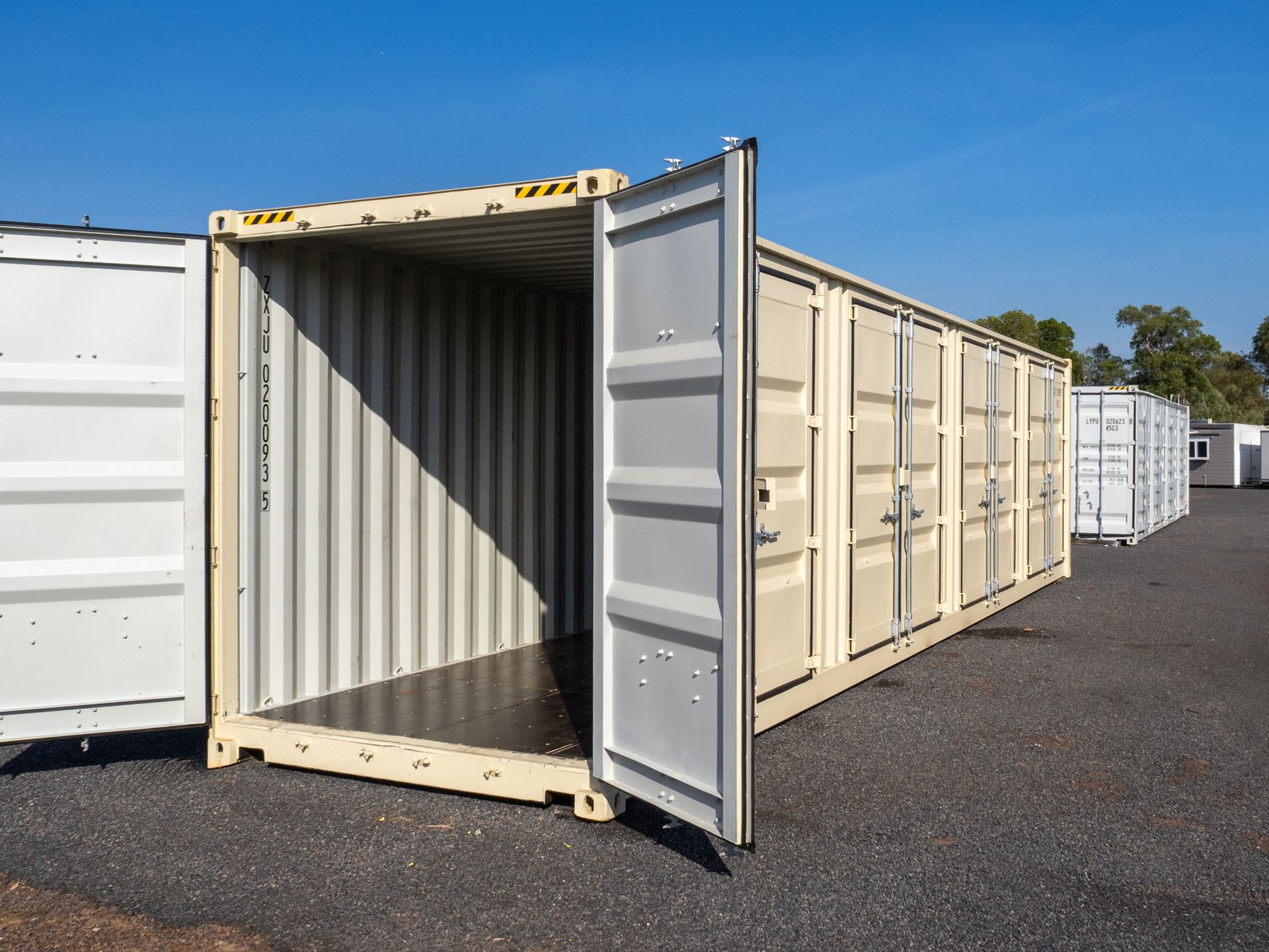 Open storage containers with beige doors, parked on gravel against a blue sky.