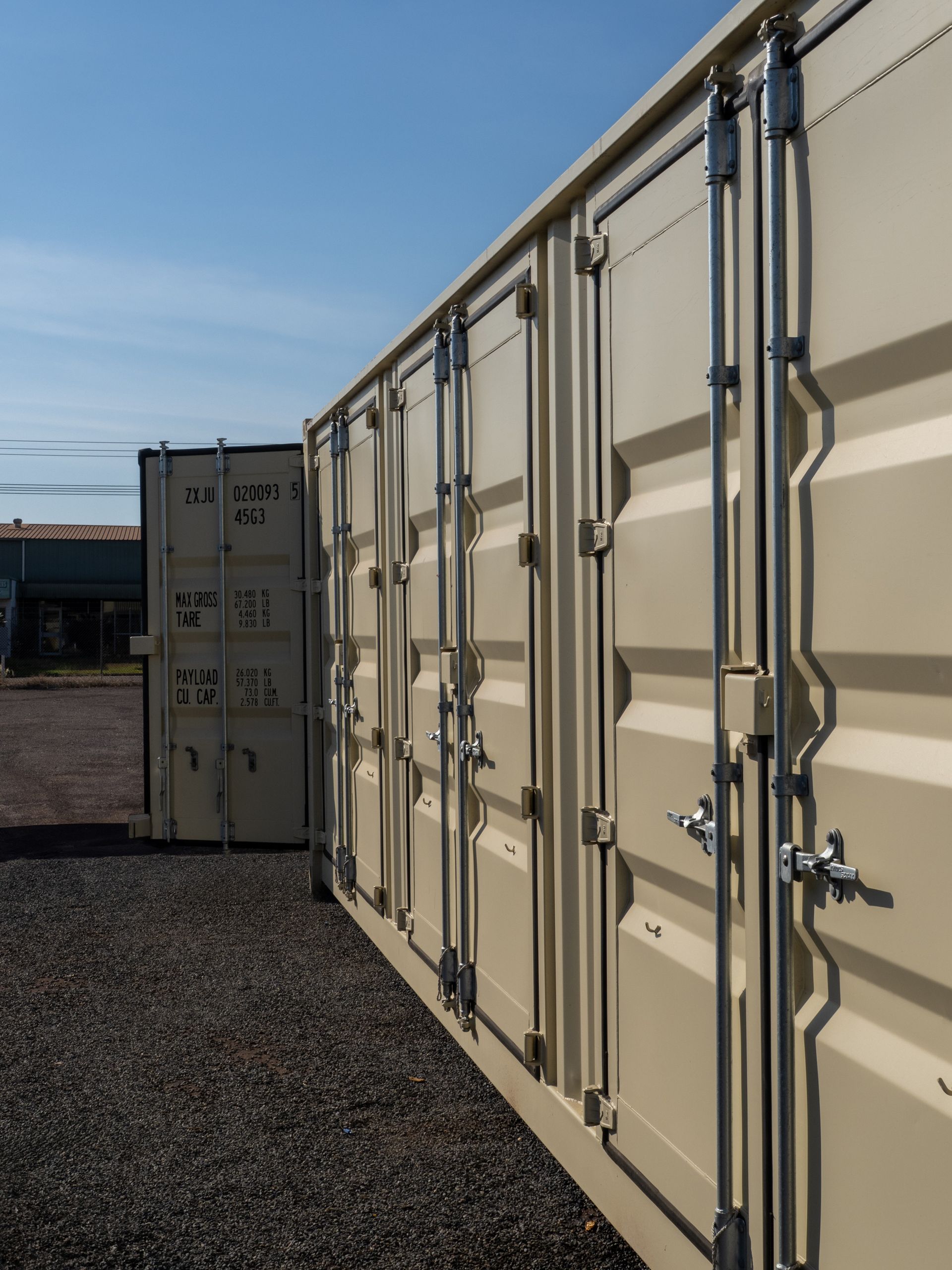 Beige shipping containers in a row, one open, on a gravel surface, under a blue sky.