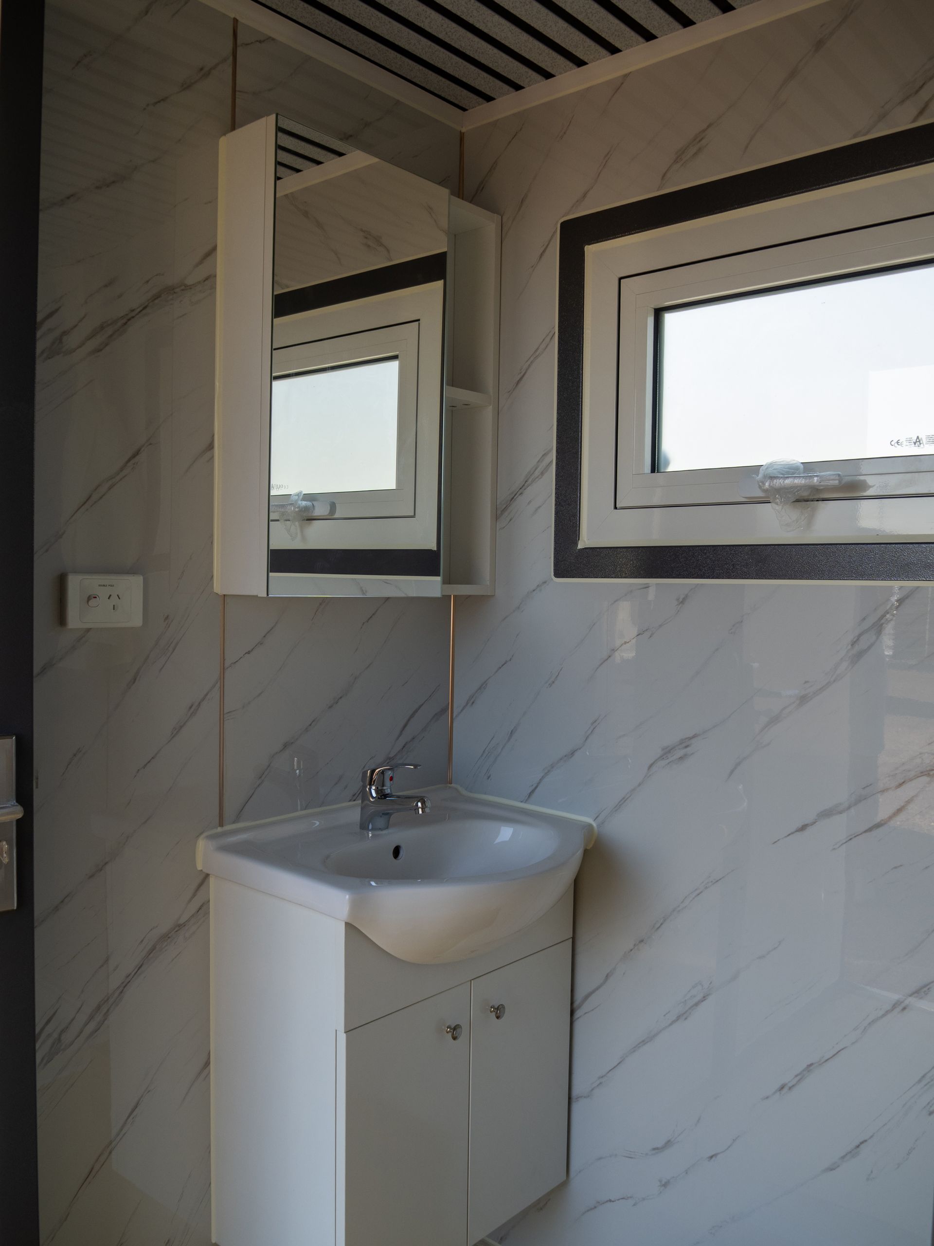 Bathroom corner with a sink, cabinet, mirror, and window, against a marble-patterned wall.