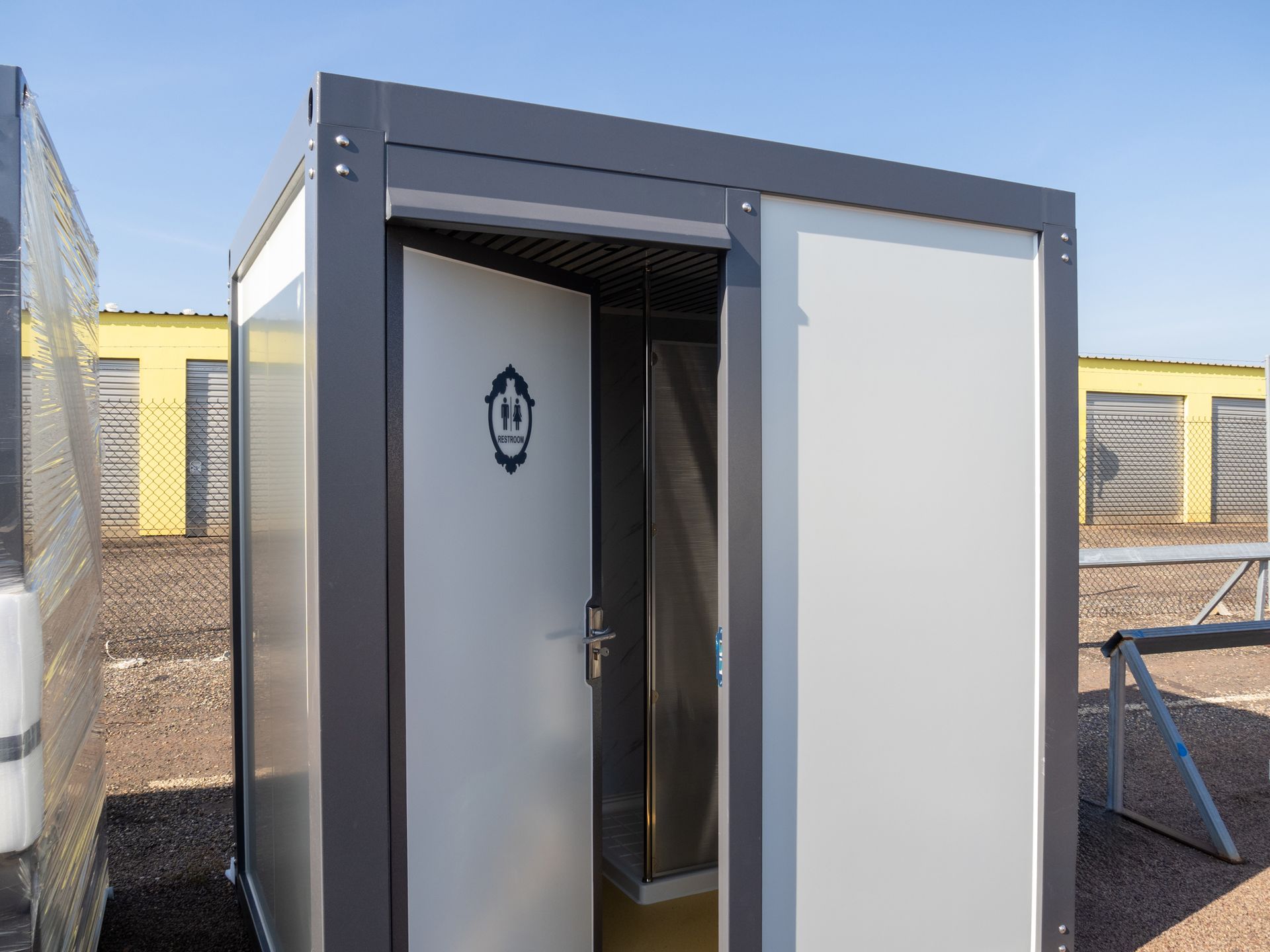 Gray metal porta-potty, open door revealing interior, set in a storage facility lot.