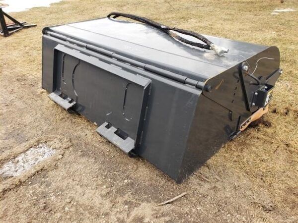 A dark gray, rectangular skid steer bucket on grass, with hydraulic lines visible on top.