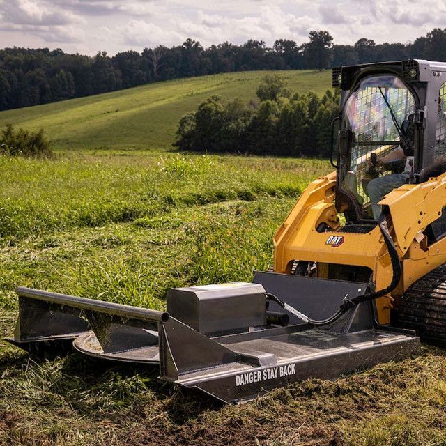Yellow Caterpillar skid steer mower cutting grass in a field with trees on a hill in the background.