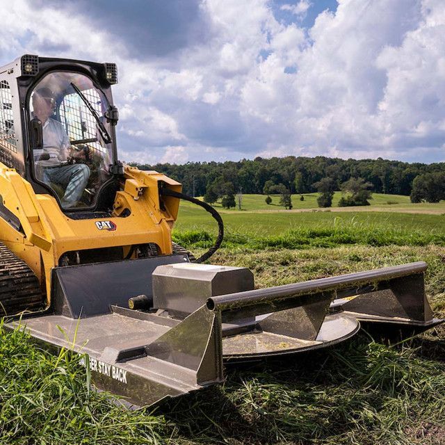 Yellow Caterpillar skid steer mower cutting grass in a field with trees on a hill in the background.