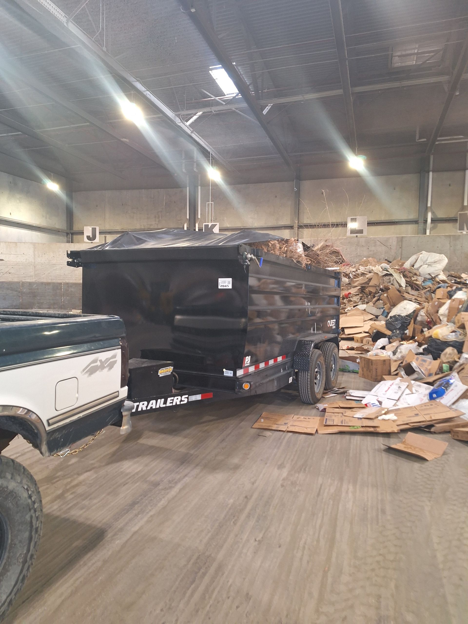 Black trailer filled with debris, hitched to a white truck inside a concrete building. Cardboard boxes and debris litter the floor.