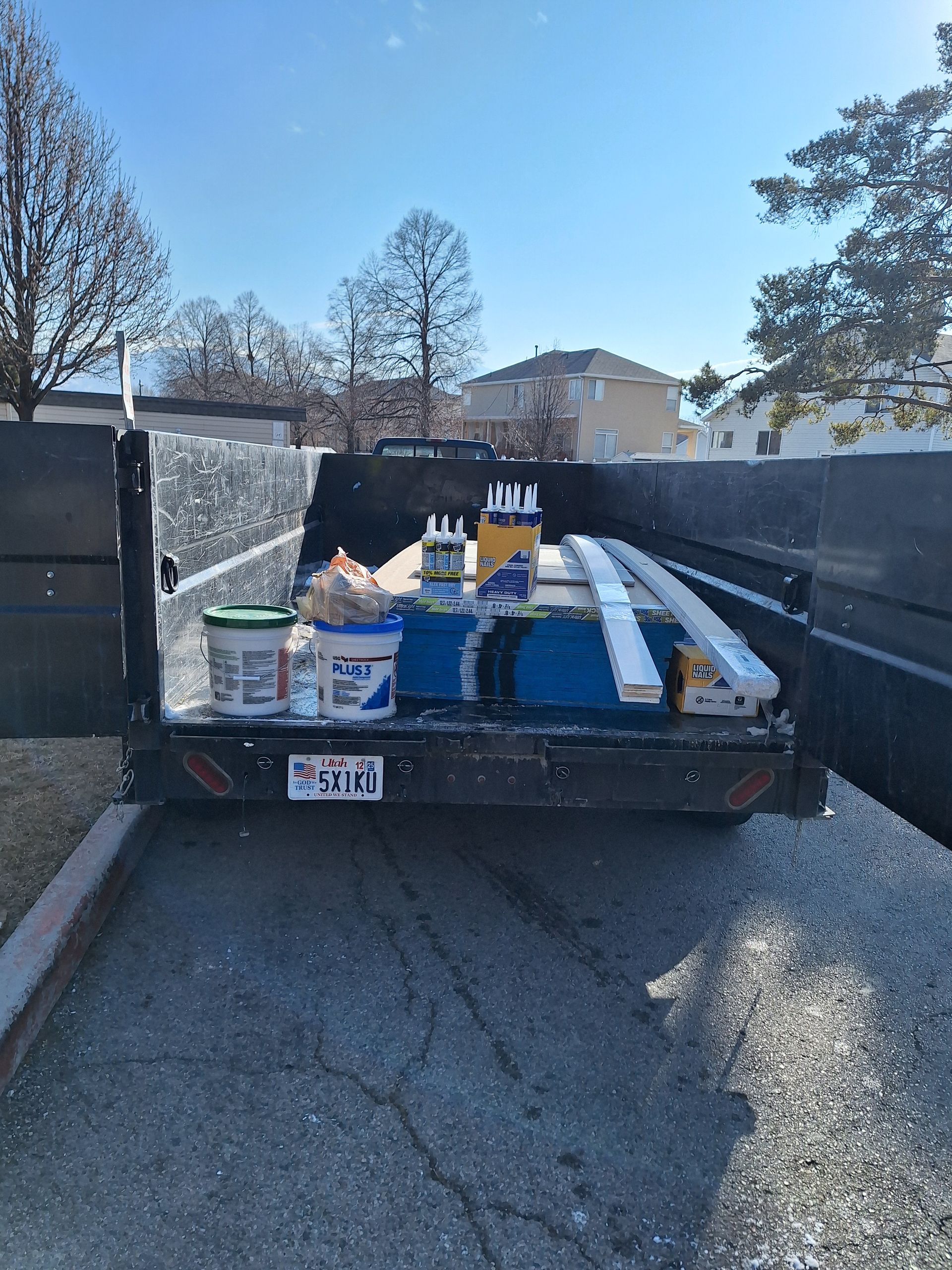 Two men loading lumber onto a red trailer in a warehouse with shelves of wood. One man wears a red shirt, and the other wears a navy shirt and jeans.