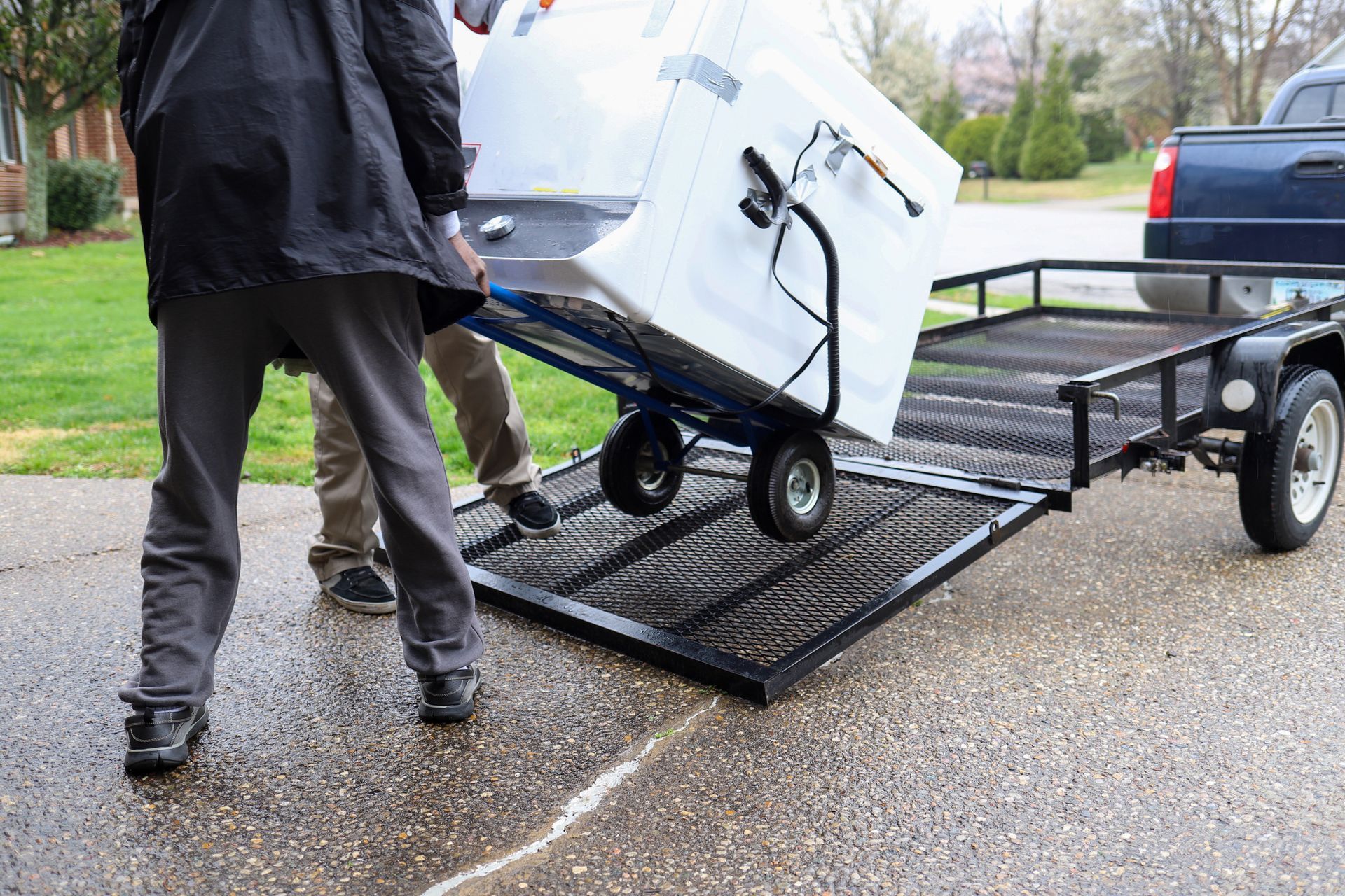 Two people loading a large appliance onto a trailer using a hand truck in a driveway.