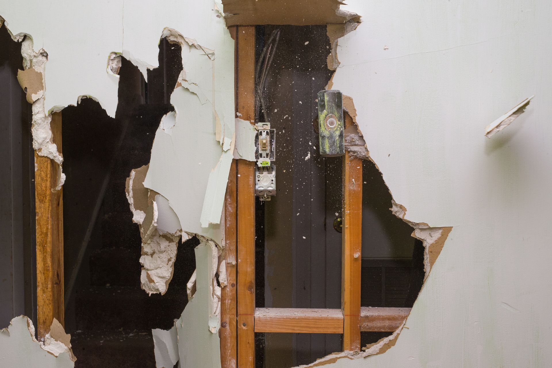 Damaged wall with a large hole exposing wooden framing and electrical components. Beige drywall is torn, and debris is scattered.