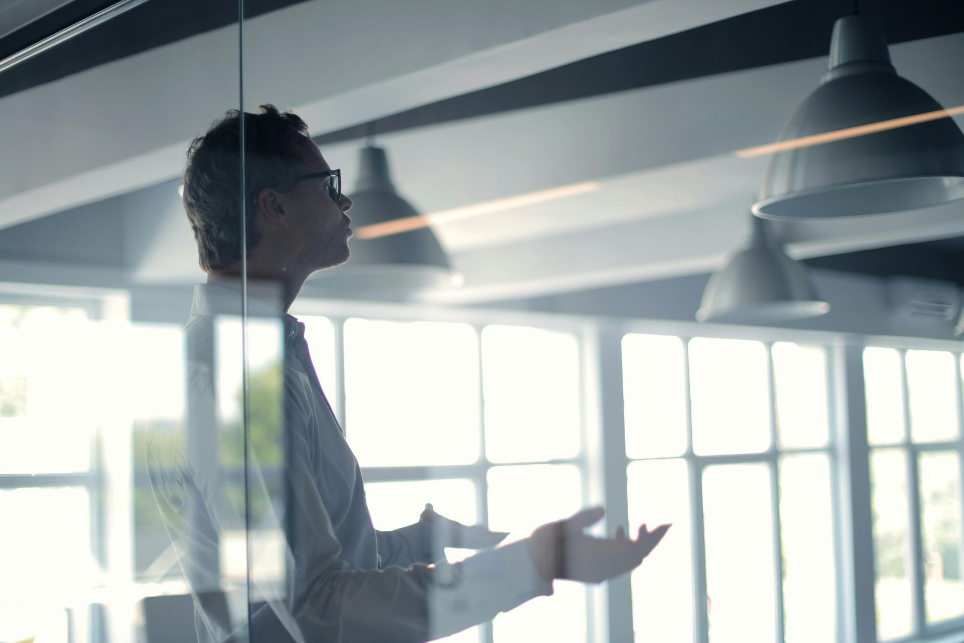 Man in glasses gestures with hands inside a modern office with overhead lights, seen through glass.