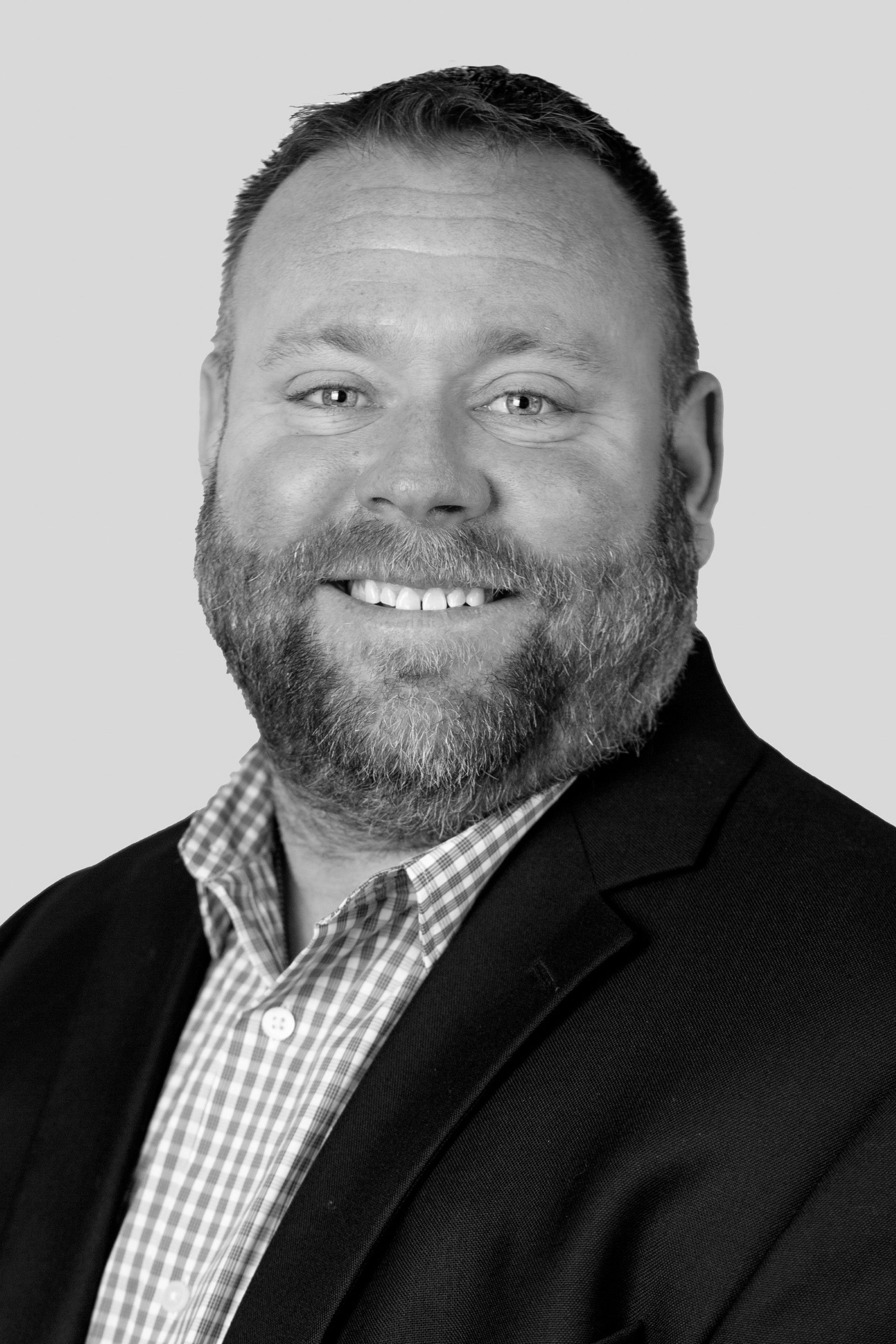 Man with beard smiling, wearing a button-down shirt and blazer, against a light gray background.