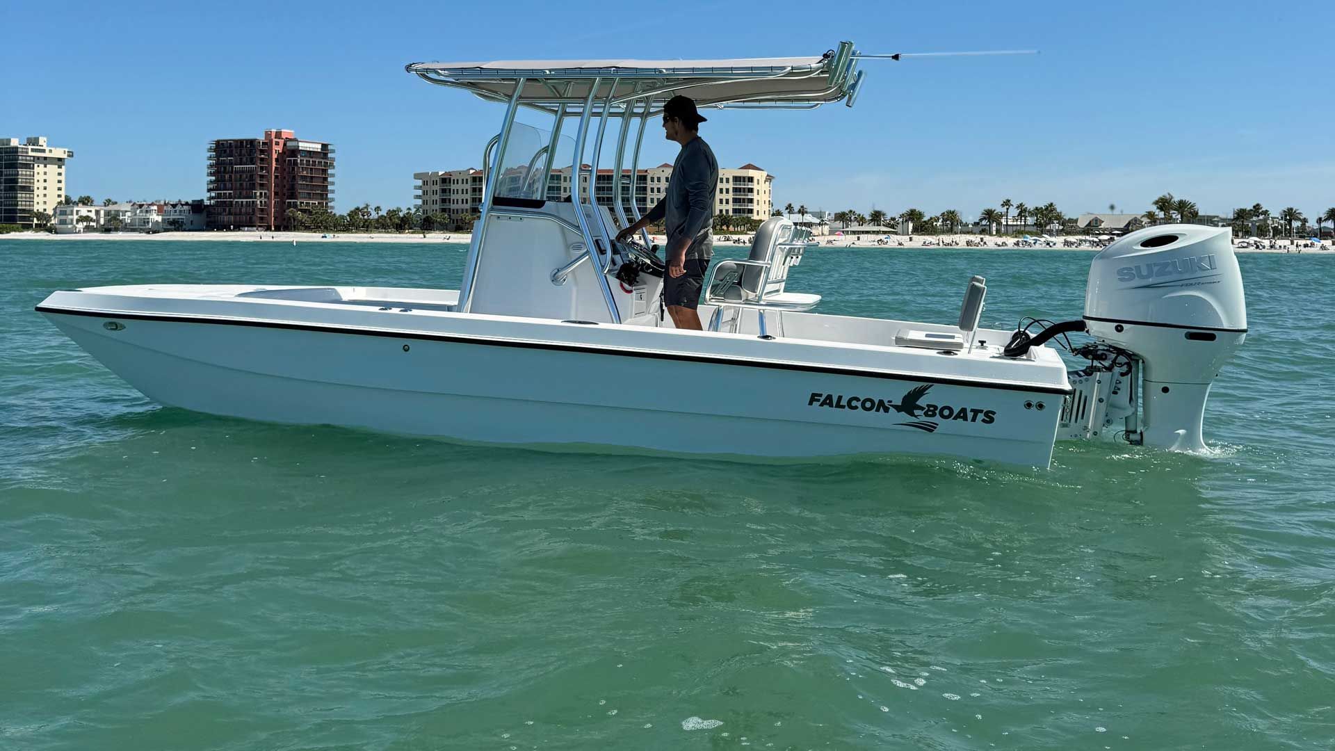 A white center console boat with a person at the helm is in shallow water. Buildings and beach are visible in the background.