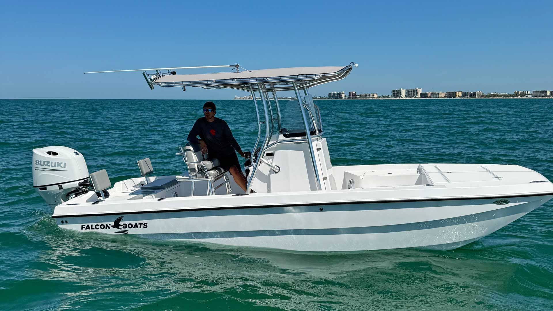 A person stands at the helm of a white center-console boat on the water. Buildings line the distant shore on a sunny day.