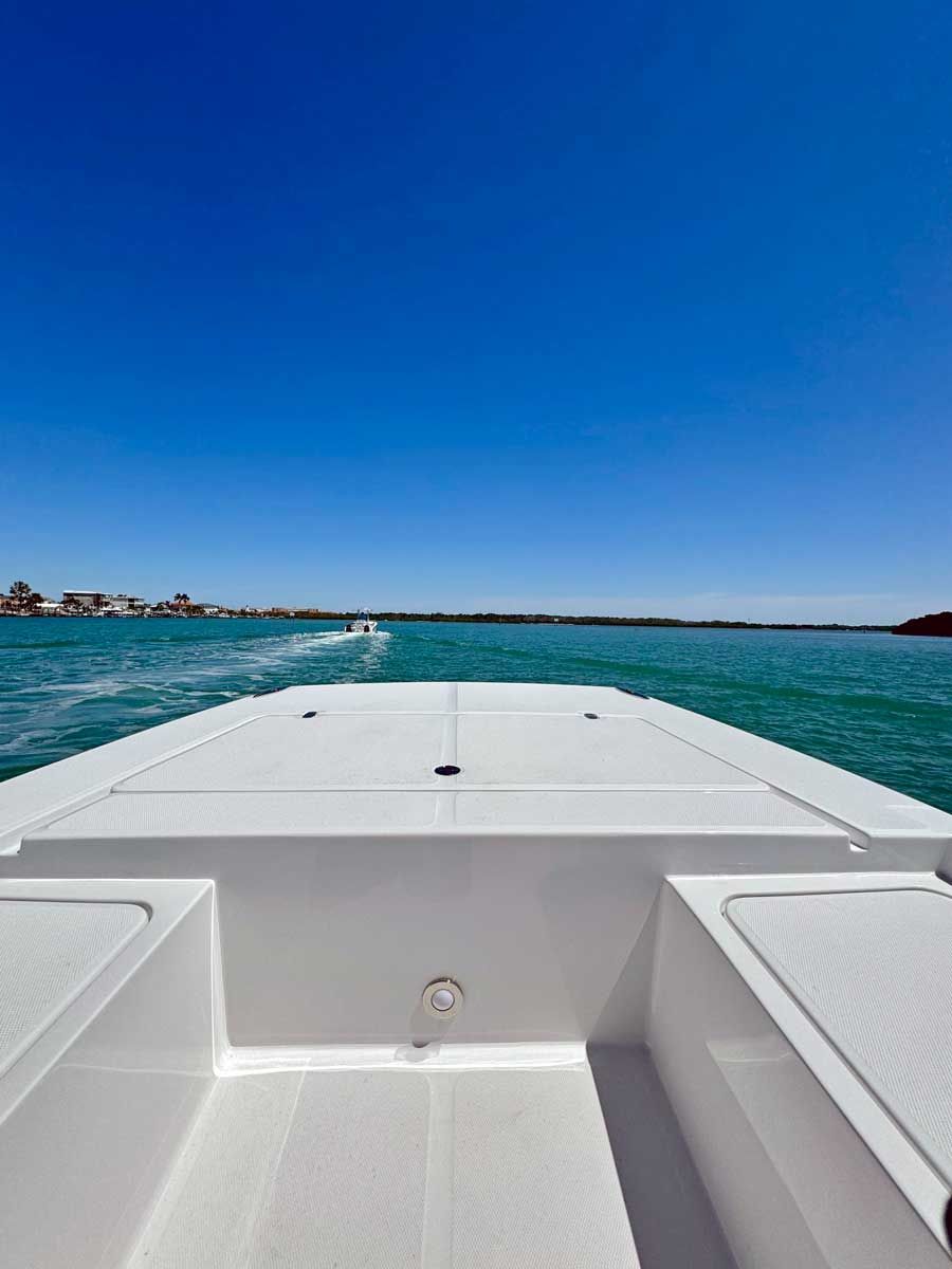 View from a white boat on turquoise water under a clear blue sky. Small wakes trail behind the boat; a shoreline is visible in the distance.