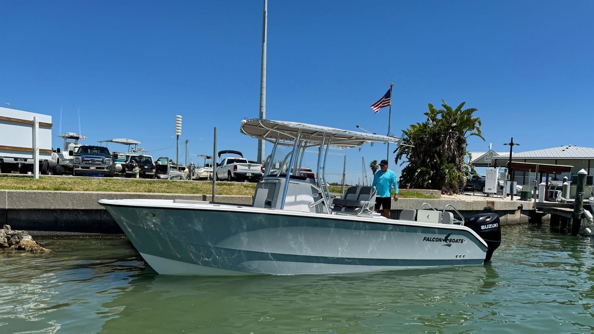 A grey and white motorboat floats in the water near a dock on a sunny day; a person stands on the boat.