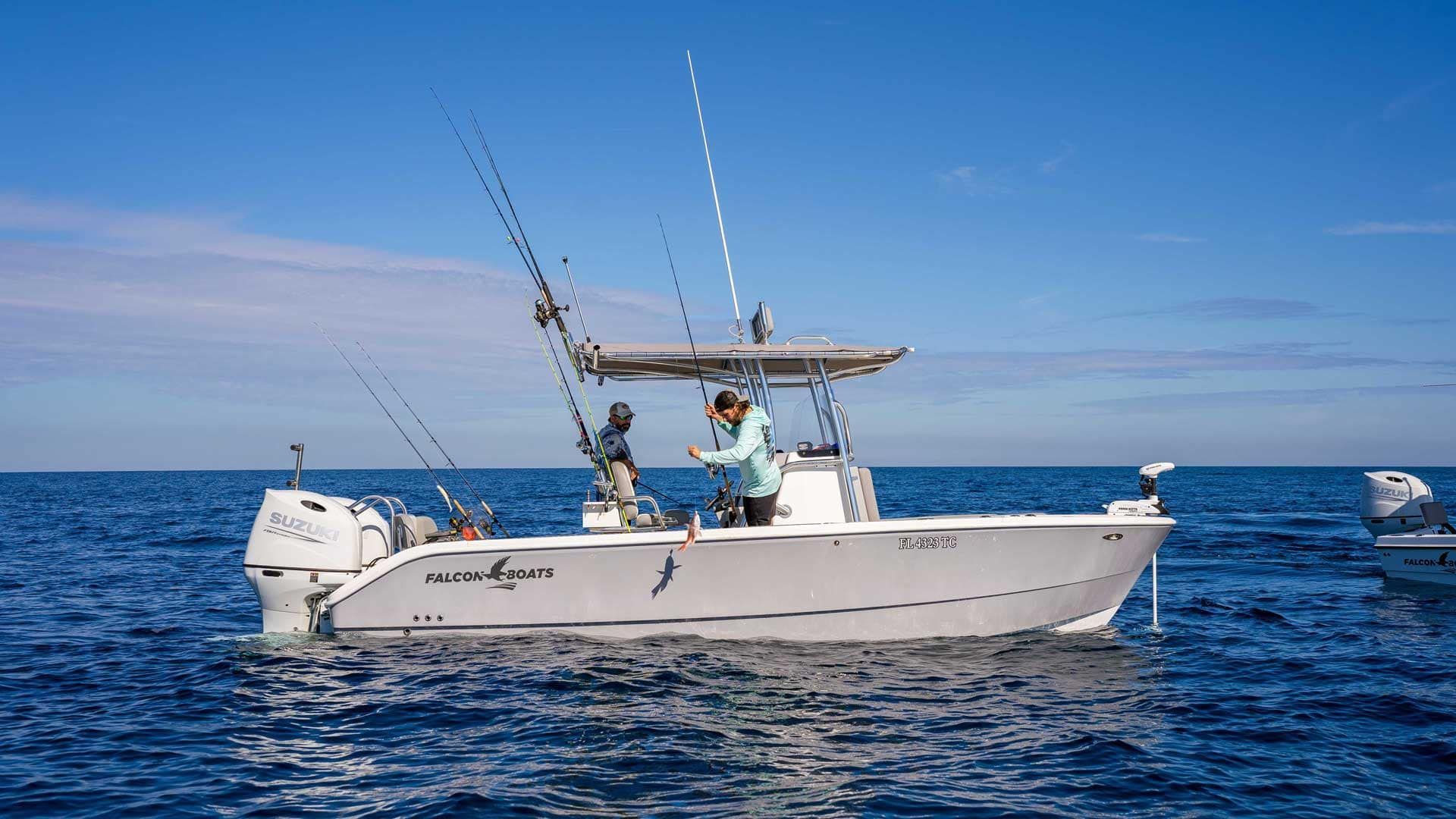 A white fishing boat on blue water with two people fishing; clear blue sky.