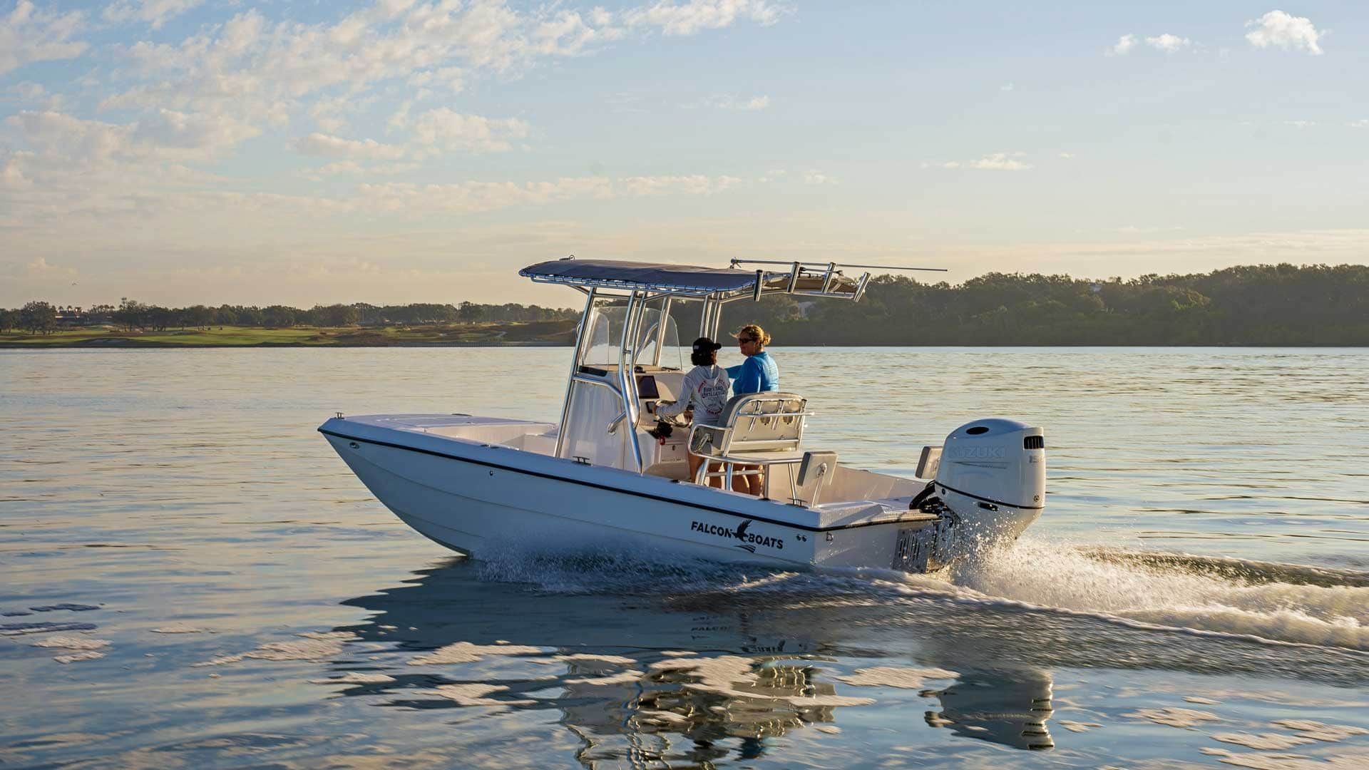 A white motorboat with two people on board speeds across calm water towards a treeline under a sunny sky.