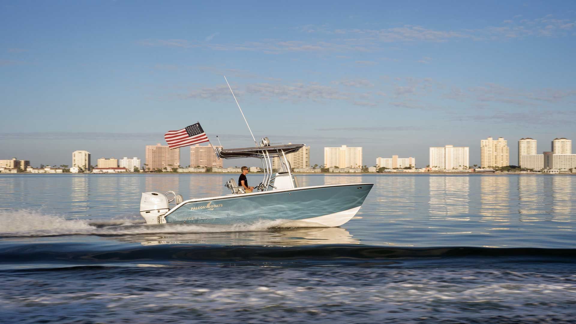 A blue and white motorboat with an American flag cruises on the water, with a city skyline in the background.