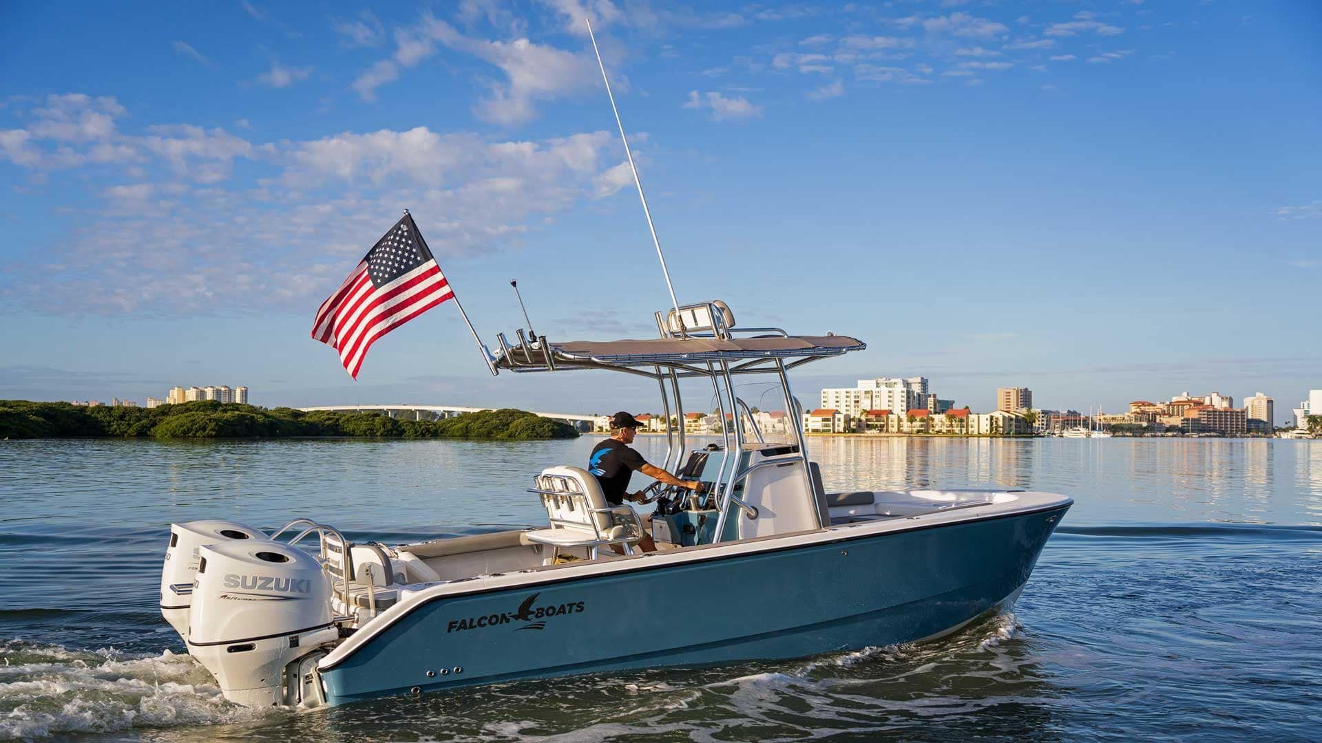 A blue center console boat with two white motors underway on water, flying an American flag. A person steers the boat under a sunny sky.