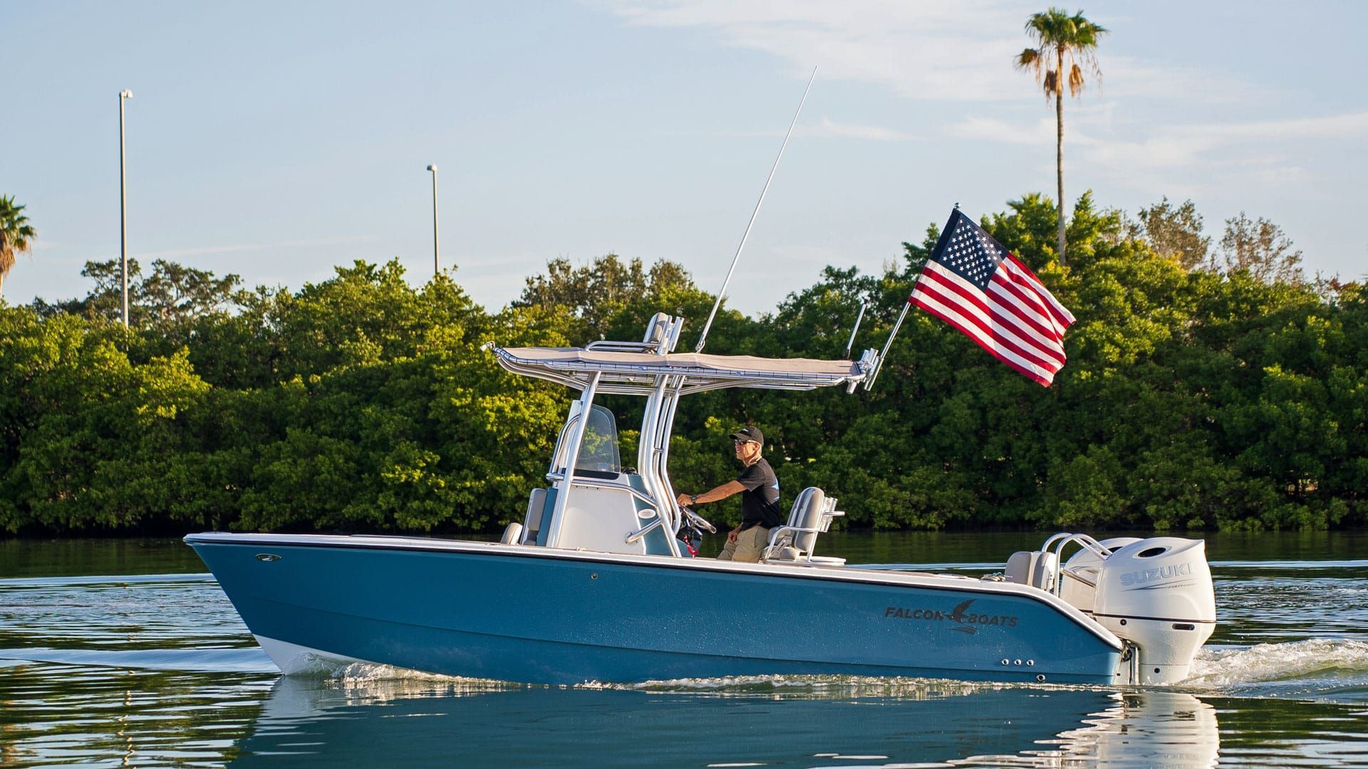 Blue motorboat with an American flag sailing on water, a person steering. Lush green foliage and palm trees are in the background.