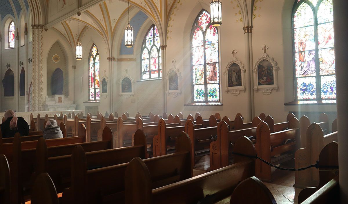 Streams of sunlight shining through stained glass windows in a Catholic church sanctuary with long wooden pews