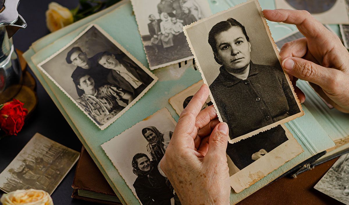 Older Hispanic woman holding at her mother’s aged black and white photo with a family photo album in the background, surrounded by flowers.