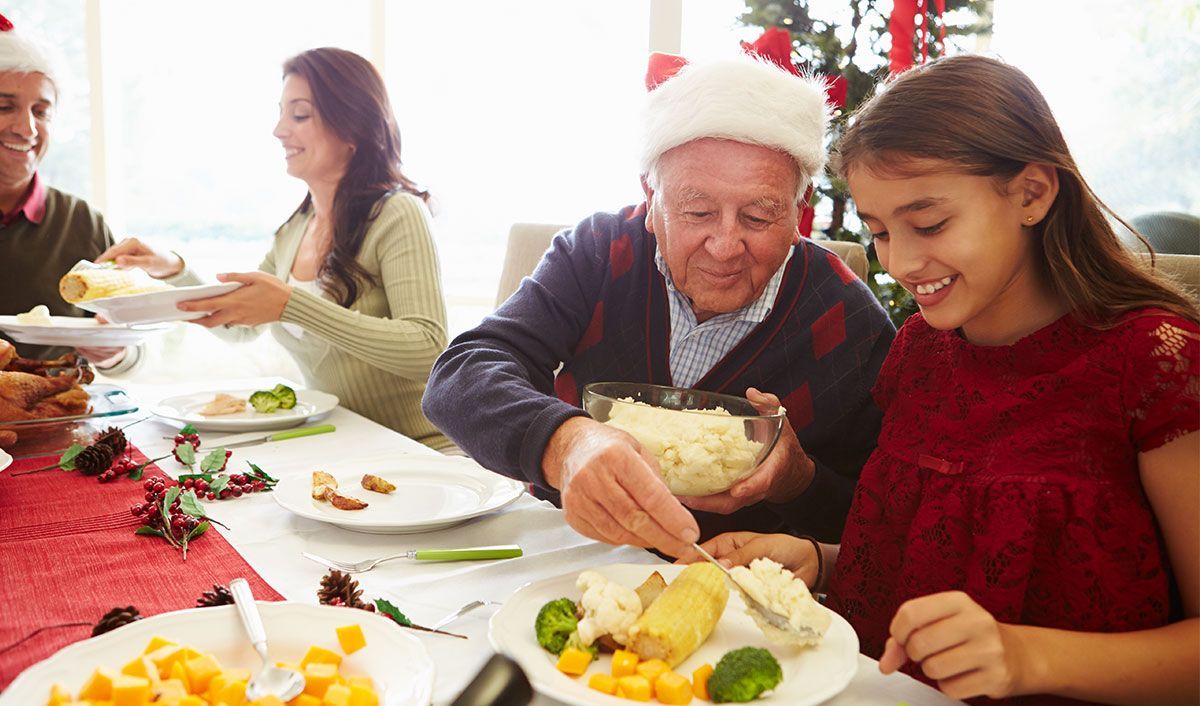 Hispanic family eating a holiday meal together while the Grandfather wearing a Santa hat serves his granddaughter mashed potatoes