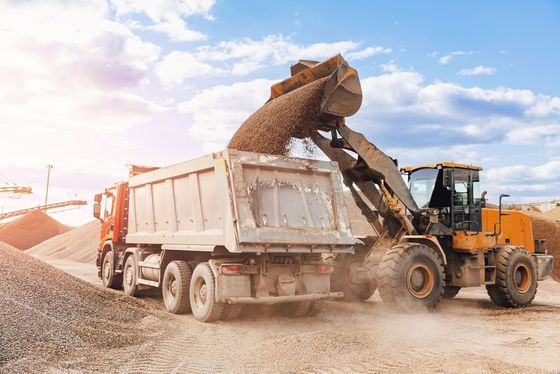 Loader dumping gravel into a white dump truck at a construction site, with dust and blue sky.