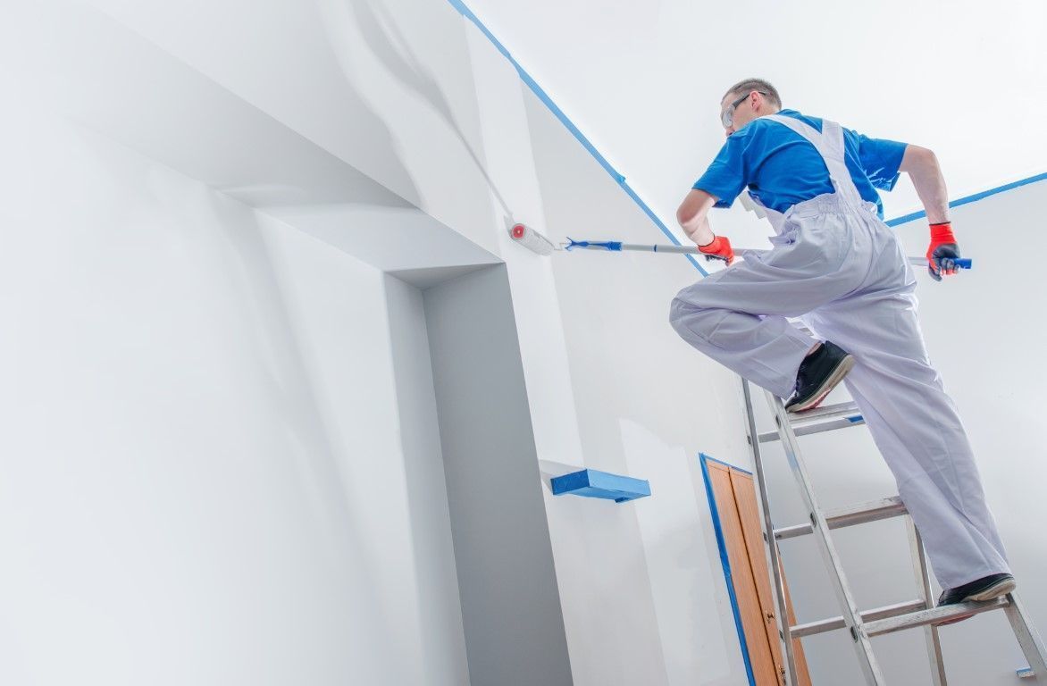 Painter on a ladder applying white paint to a high ceiling corner.