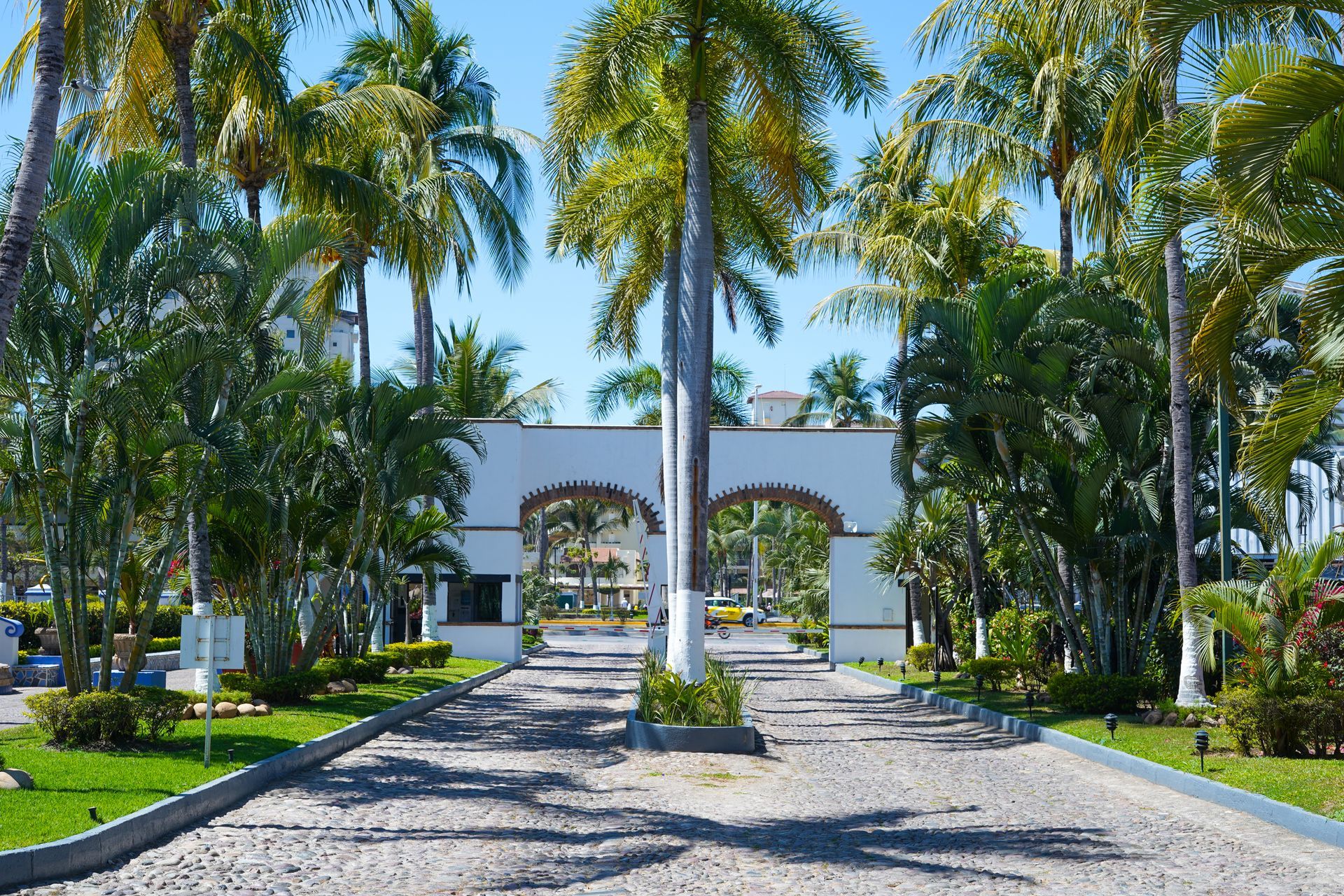 A gated entrance with white arches, palm trees, and a cobblestone driveway leading into a resort.