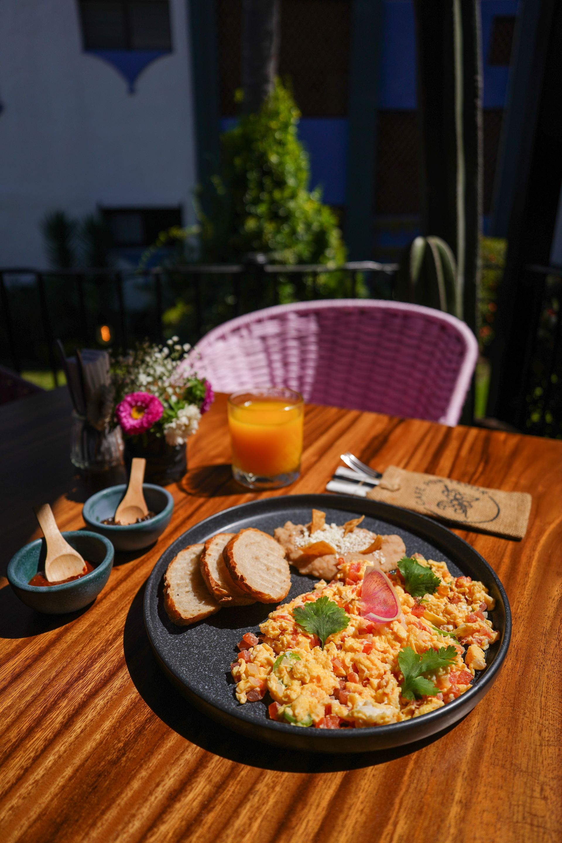 Scrambled eggs, toast, and juice on a wooden table outdoors. Pink wicker chair and colorful building in the background.