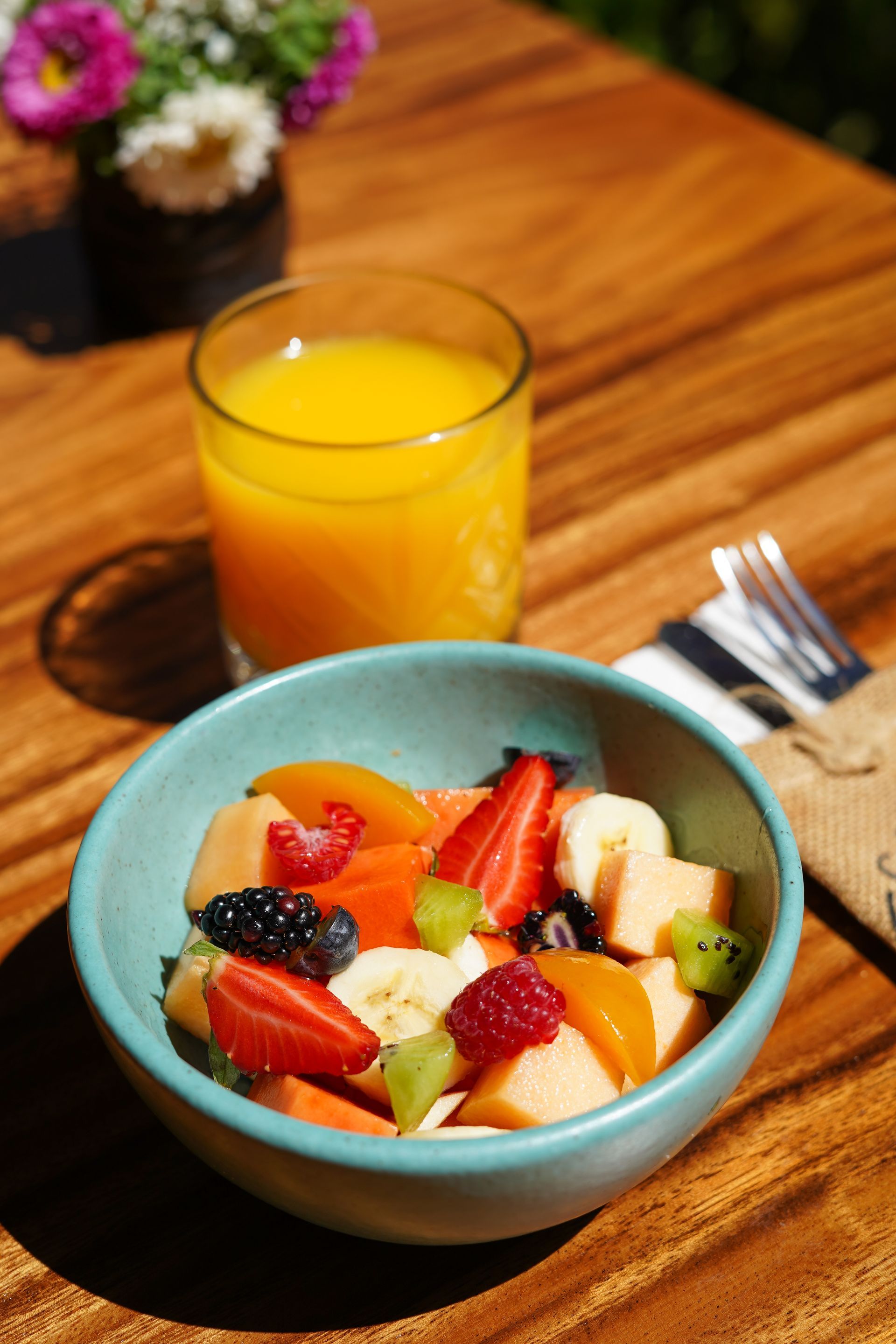 Fruit salad in blue bowl, orange juice, and a small flower arrangement on wooden table.