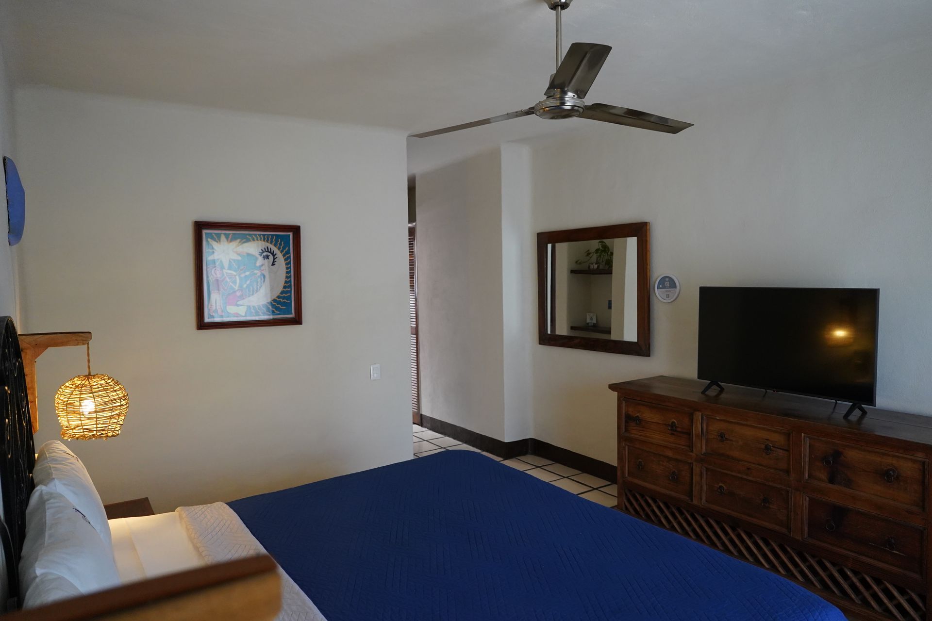 Bedroom with blue bedspread, dresser, TV, mirror, ceiling fan, and art.