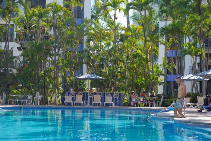 Swimming pool with a man standing at the edge. People sit at a poolside bar, surrounded by palm trees.