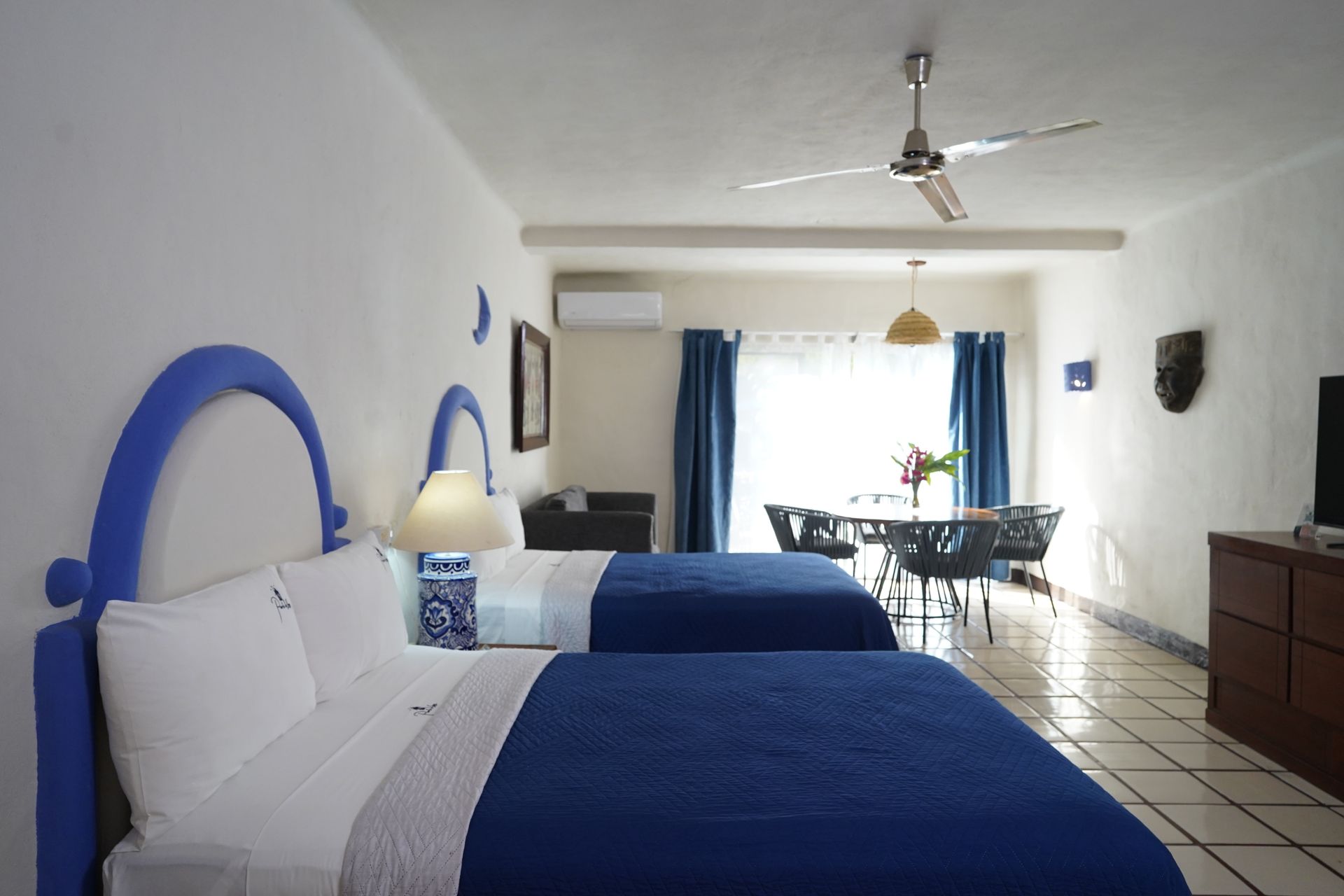 Two beds in a bright room, blue accents on headboards, matching bedspreads, dining area with table and chairs.